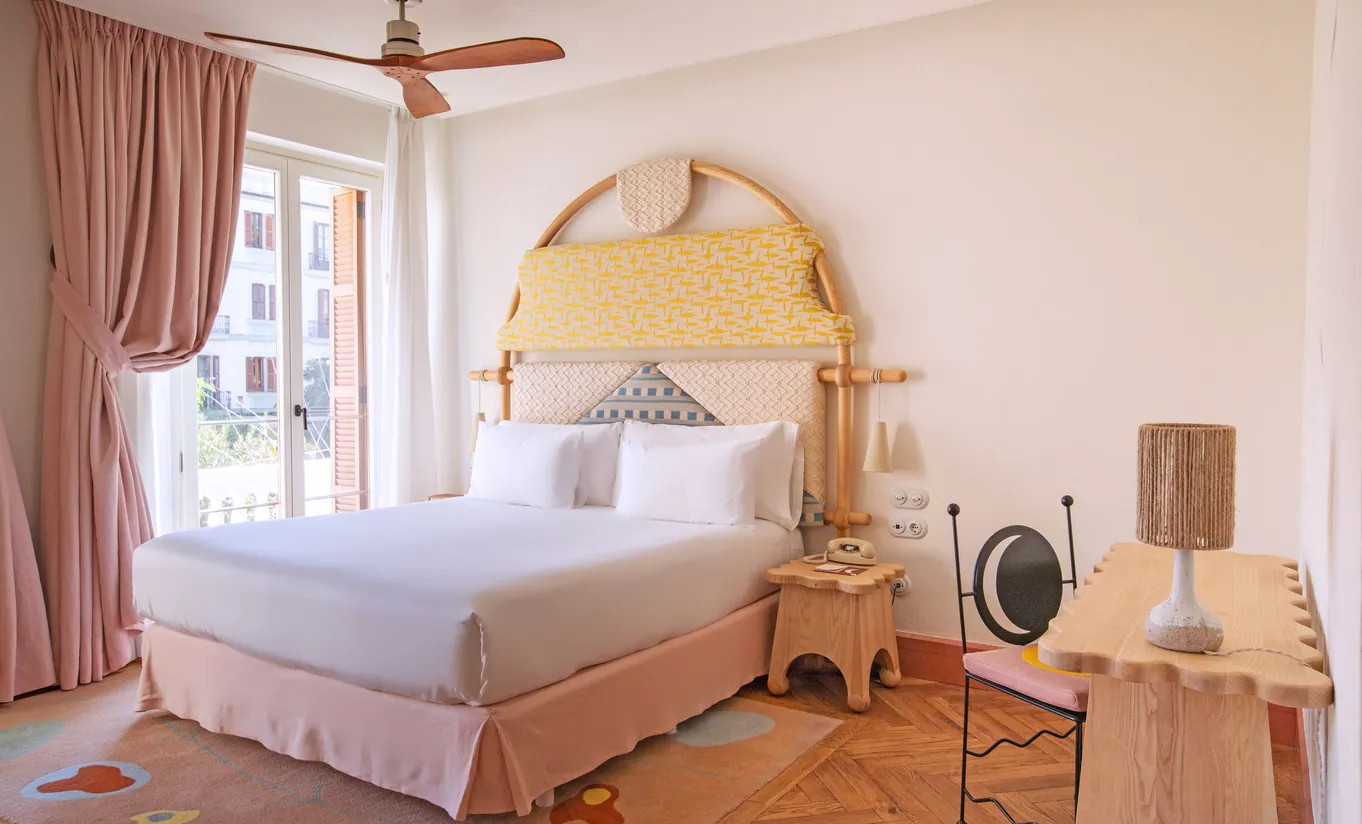 Sun-filled bedroom with a soft golden arched headboard, blush pink curtains, wicker accessories and ceiling fan at Montesol Experimental hotel, Experimental Group.