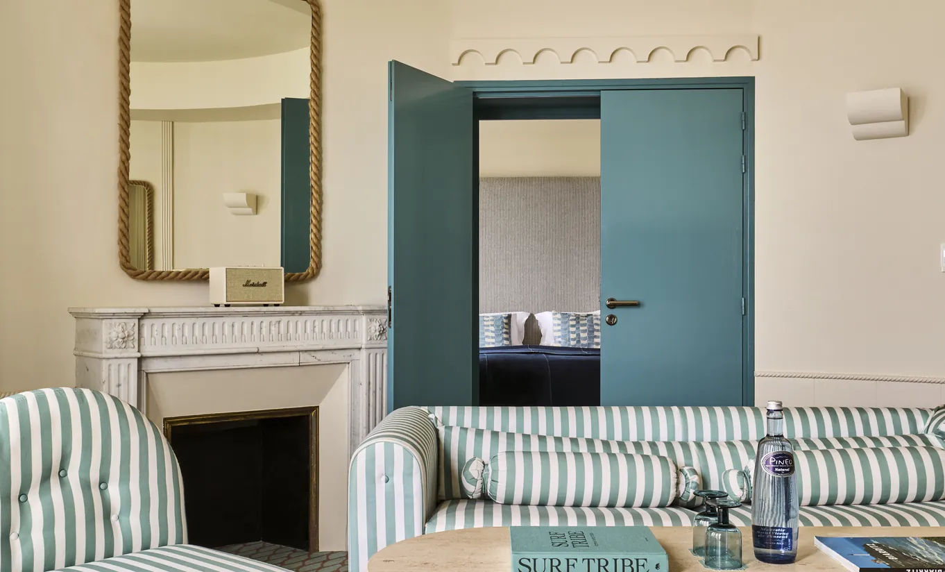 Experimental Group hotel living room with striped sofa, white fireplace, gold mirror, and teal blue arched doorway leading to bedroom