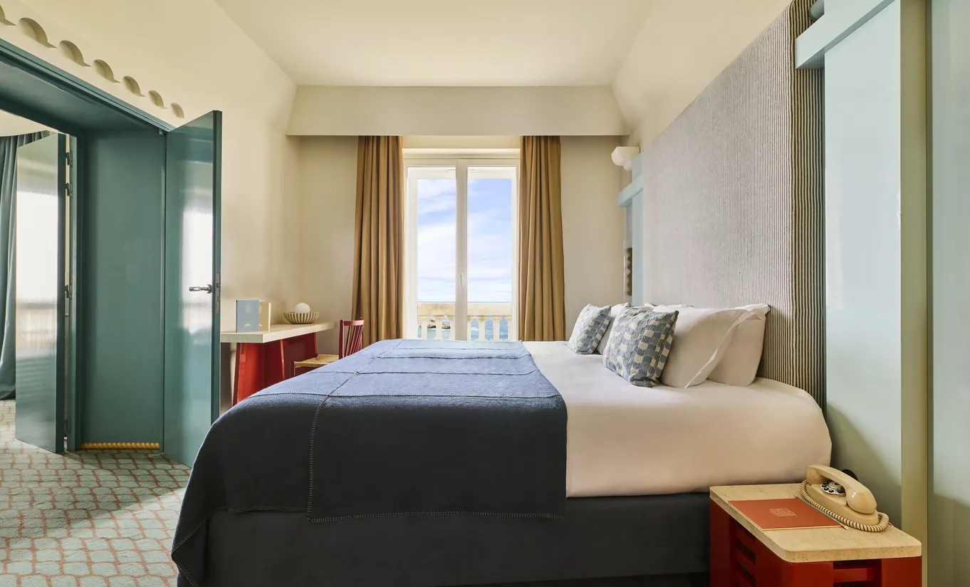 Experimental Group hotel standard room with white bedding, navy blue throw, teal green wardrobe, and bright window with city view