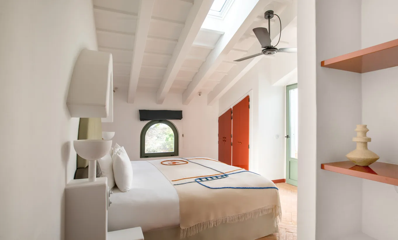 Menorca Experimental by Experimental Group – sun-drenched bedroom with exposed white timber beams, arched window and warm terracotta accents