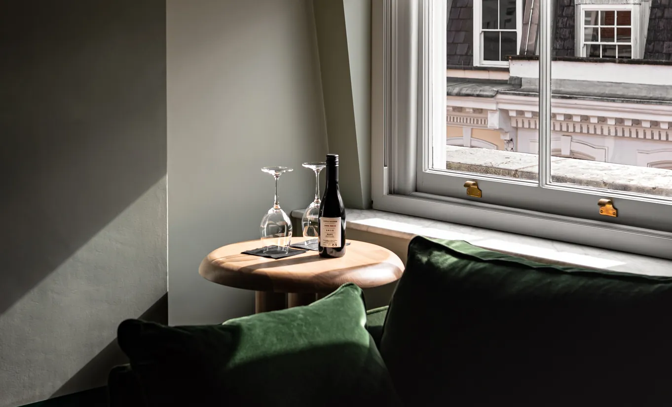 Henrietta Experimental hotel room corner with a round wooden side table, wine bottle and glasses beside a large window overlooking London