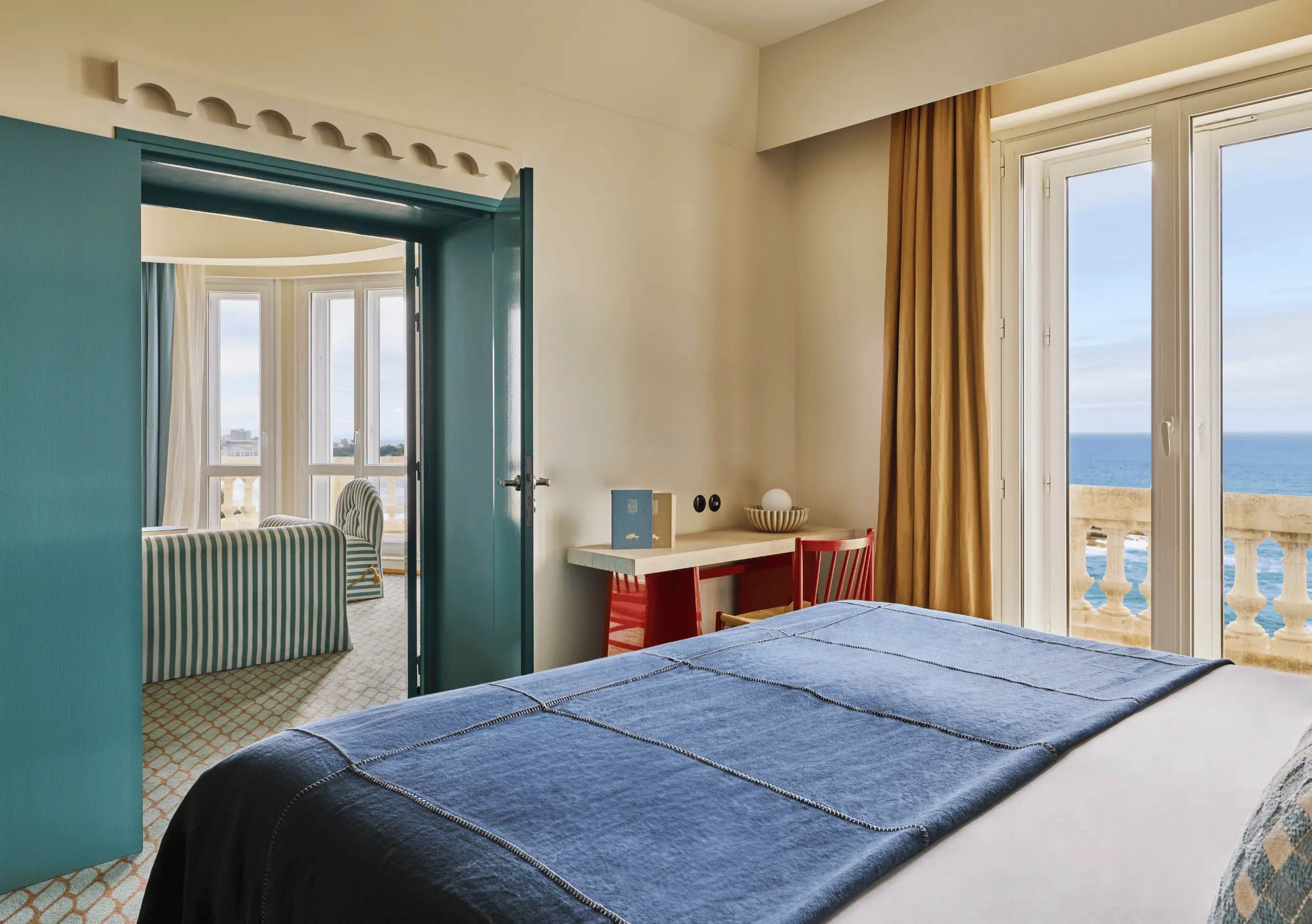 Experimental Group hotel bedroom with blue headboard, white bedding, teal bathroom door ajar, and large window overlooking the sea.