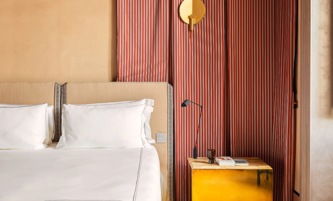 Warm hotel bedroom with white linen bedding, ochre wood-panelled wall and a sculptural yellow globe wall lamp at Experimental Roma, Experimental Group.