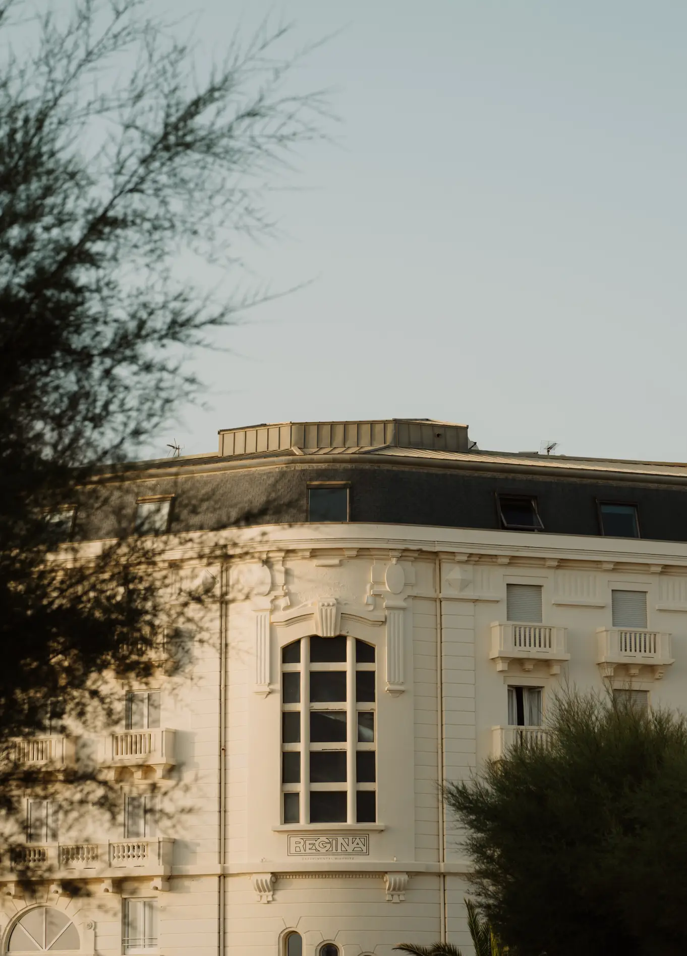 Vista parcial do exterior de um edifício clássico, com paredes claras, apresentando uma grande janela em arco, parcialmente encoberta por uma árvore em primeiro plano.