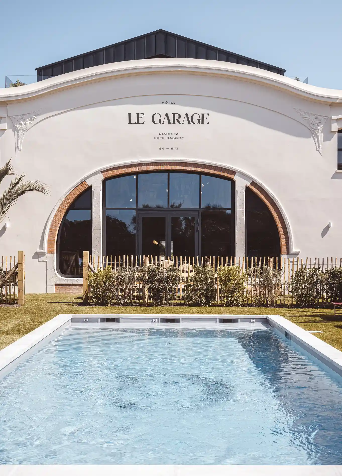 Fachada do edifício "Le Garage" com uma grande janela em arco e uma piscina em primeiro plano.