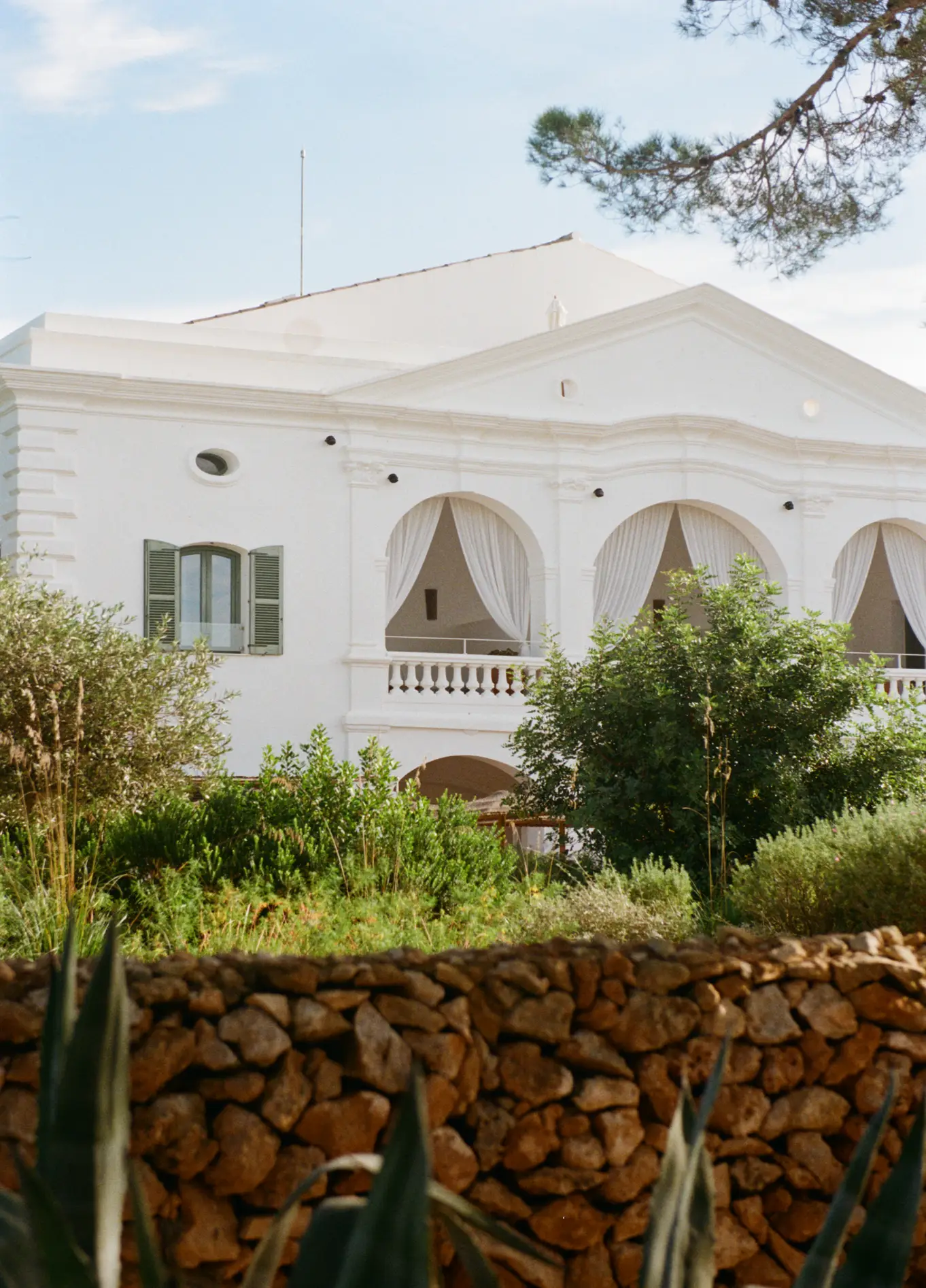 Fachada imaculadamente branca do hotel Menorca Experimental, rodeada por vegetação exuberante e um muro de pedra seca em primeiro plano.
