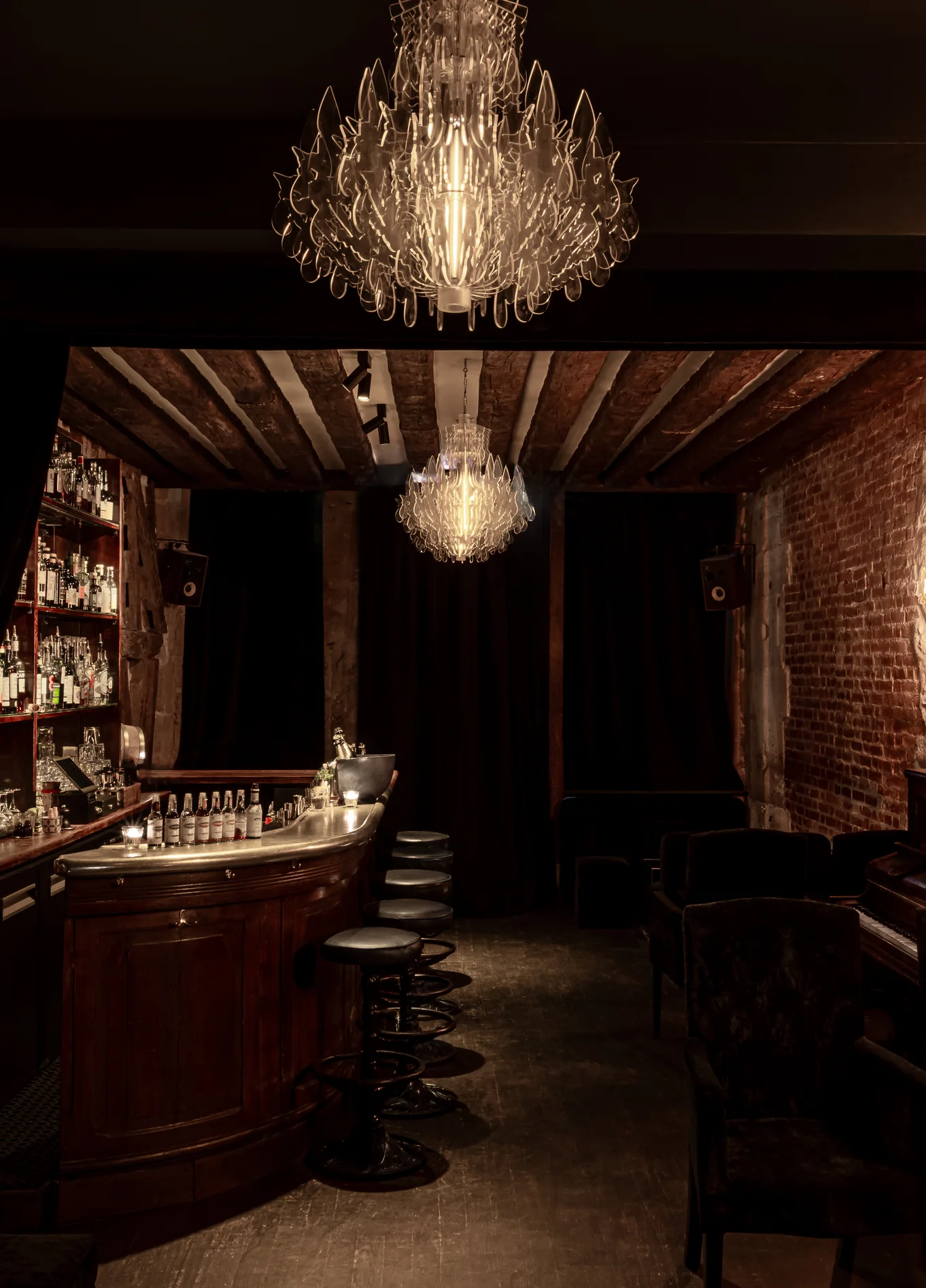 Ambiente acolhedor do Experimental Cocktail Club, com um balcão de bar em madeira, bancos corridos e um candelabro decorativo pendurado no teto.