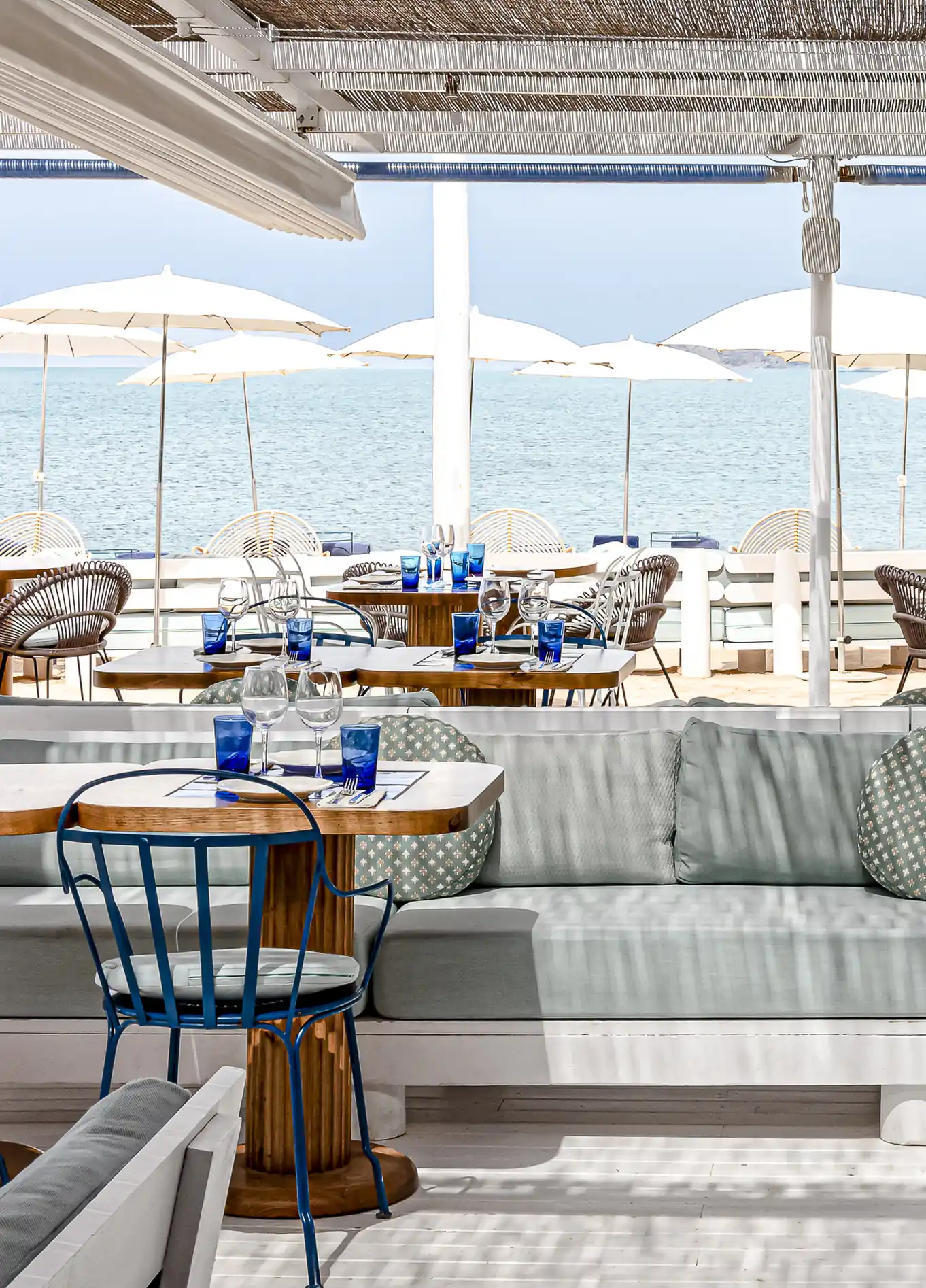 Um terraço de restaurante com sofás e mesas confortáveis, com vista para o mar e muitos chapéus de sol brancos.