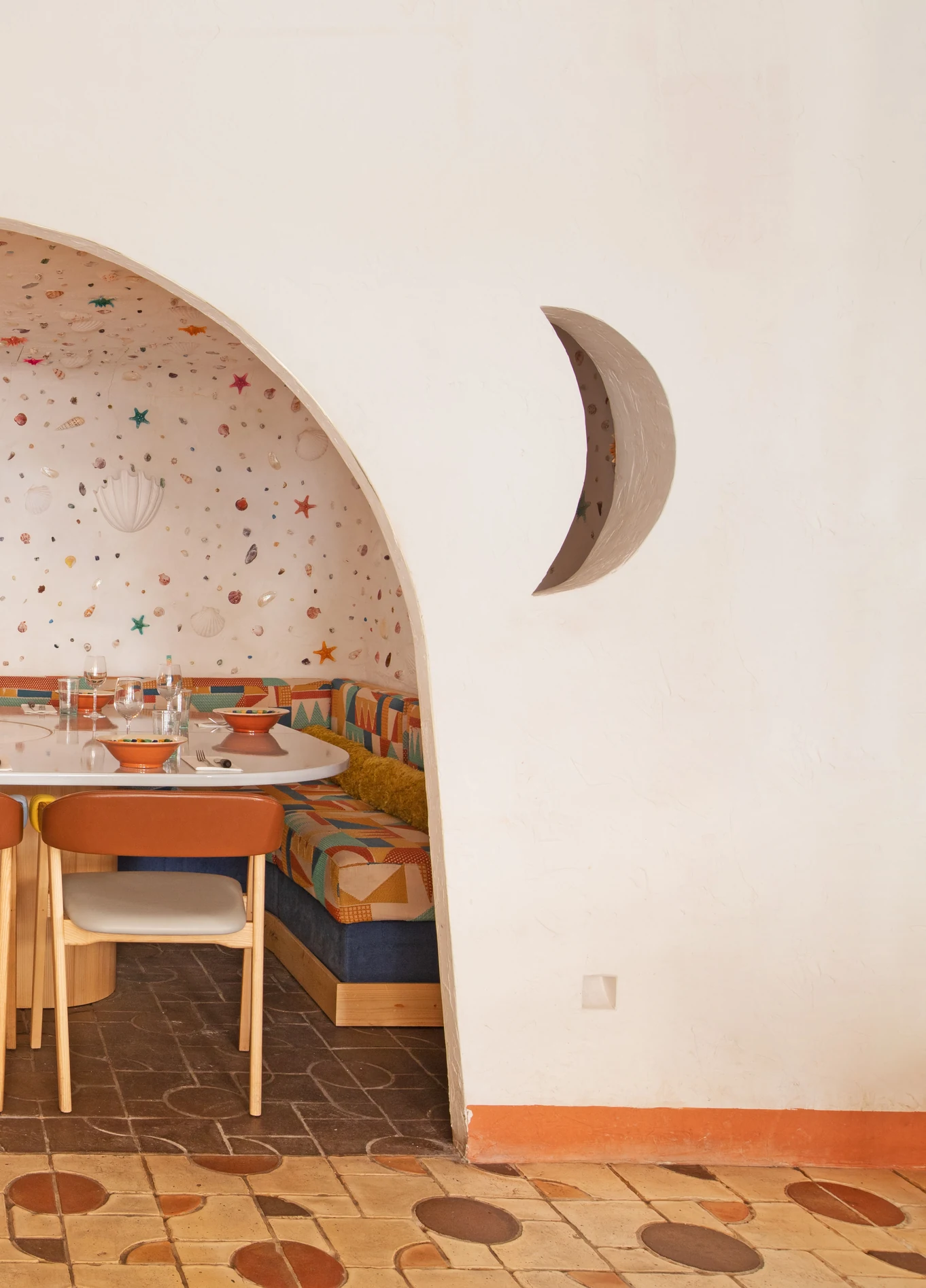 Uma área de refeições vista através de um arco, com uma decoração de parede em forma de lua, mesas com cadeiras e um chão de azulejos.