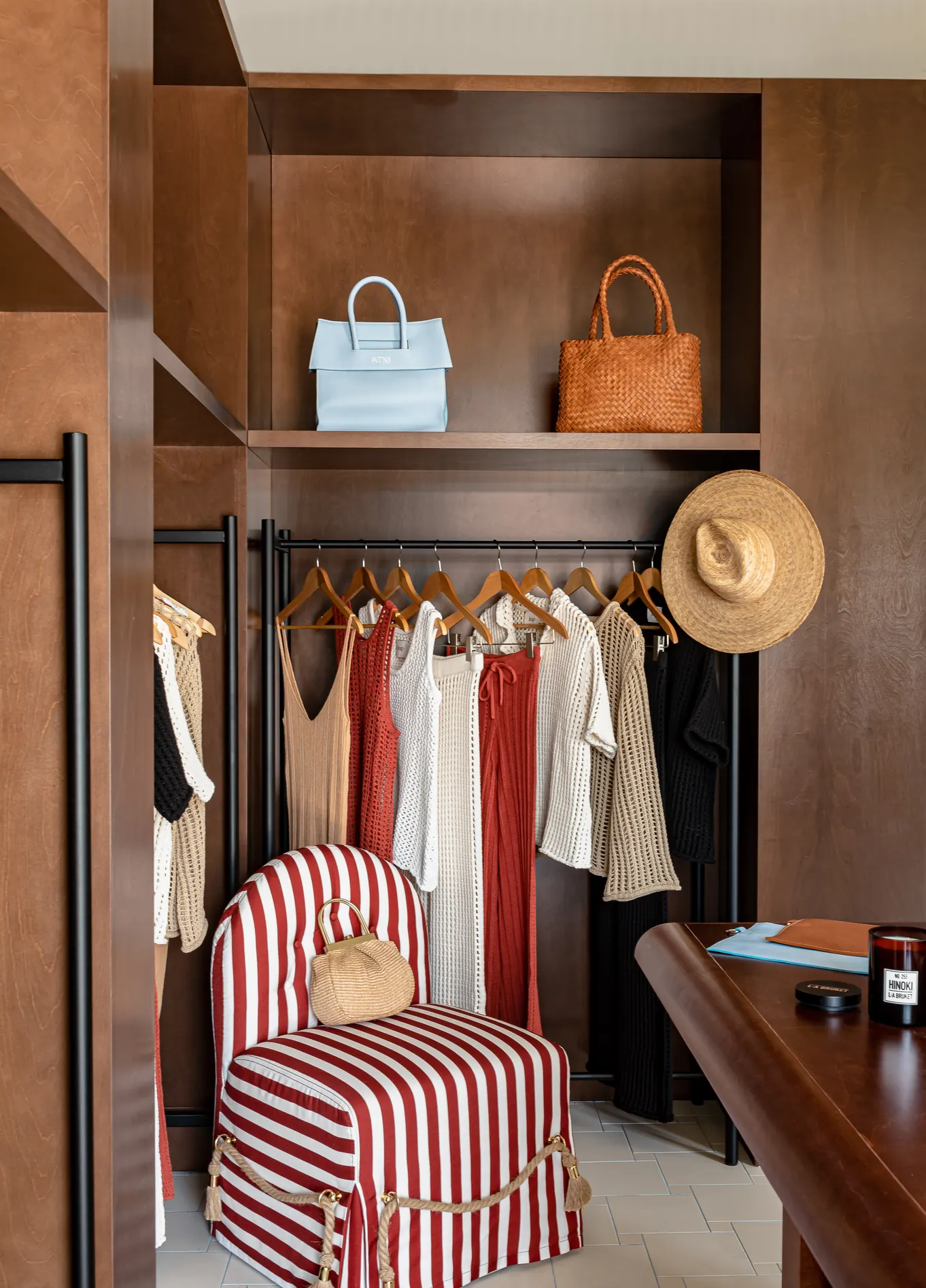 Closet walk-in elegante com roupas penduradas, bolsas na prateleira superior e uma poltrona listrada em vermelho e branco, criando uma atmosfera sofisticada.