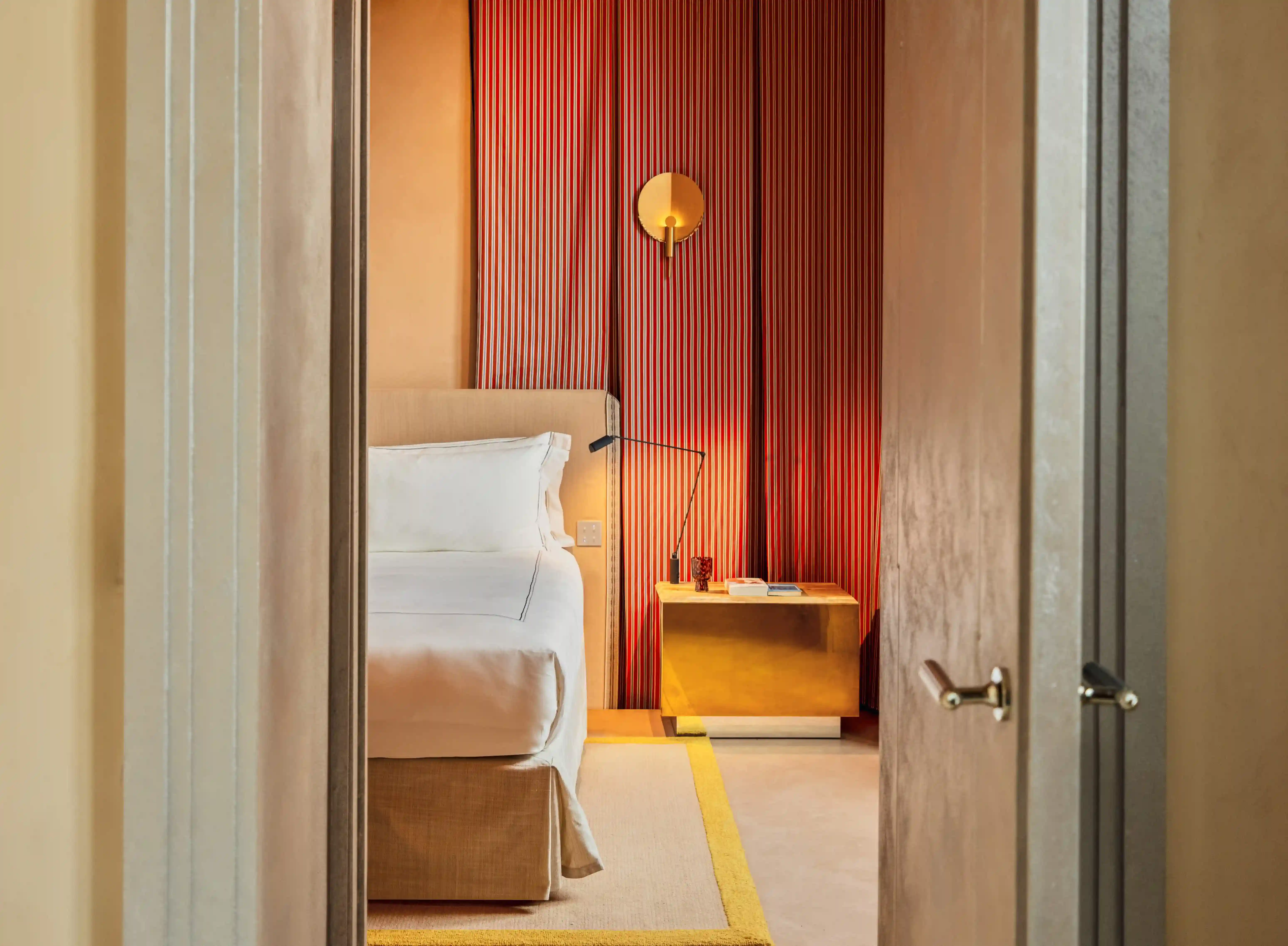 Vue encadrée, depuis l'encadrement d'une porte, d'une chambre d'hôtel comprenant un lit et une table de chevet dorée.