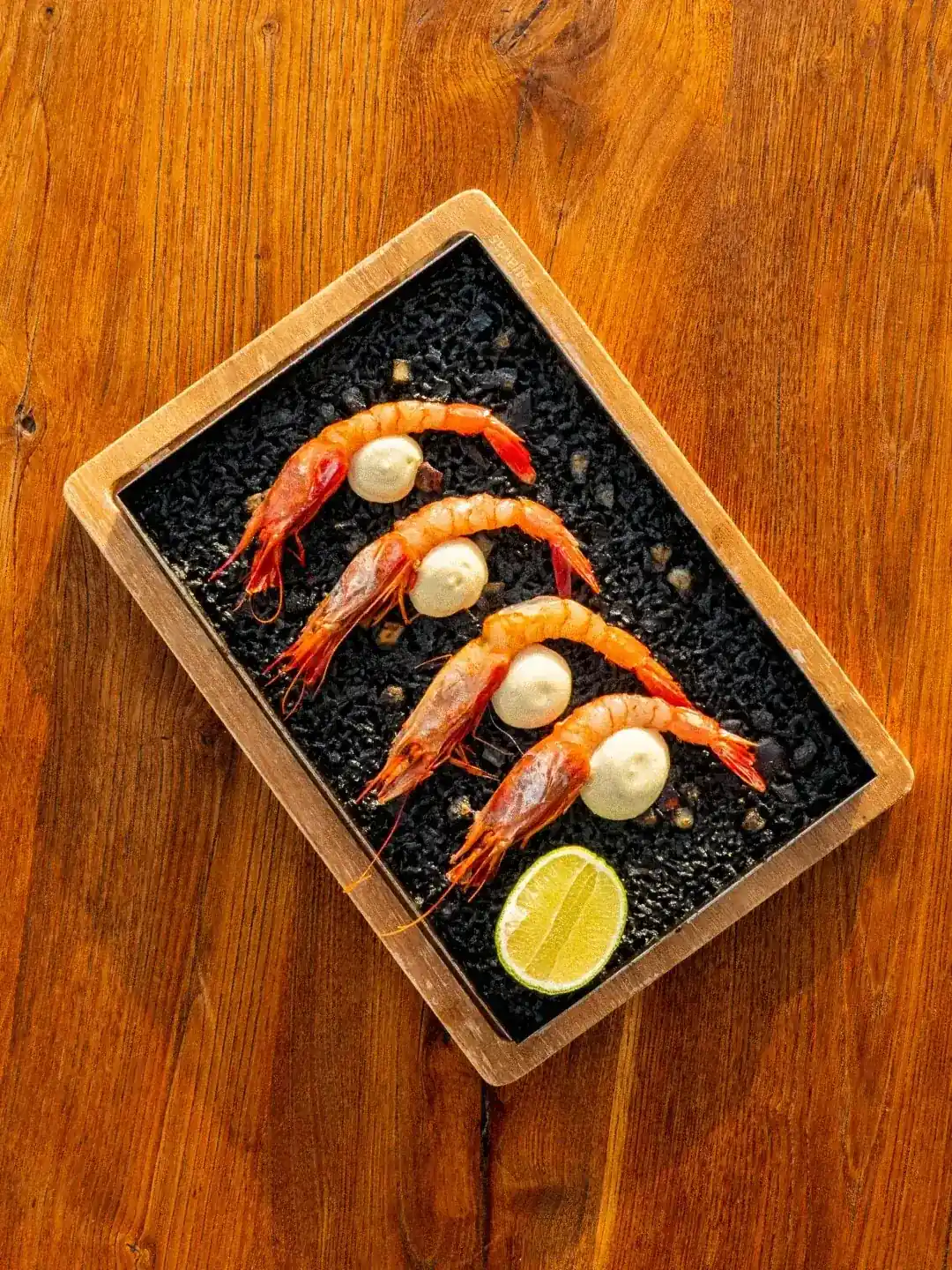 A gourmet appetizer featuring four prawns served on a dark stone slate with dollops of sauce and a lime wedge.