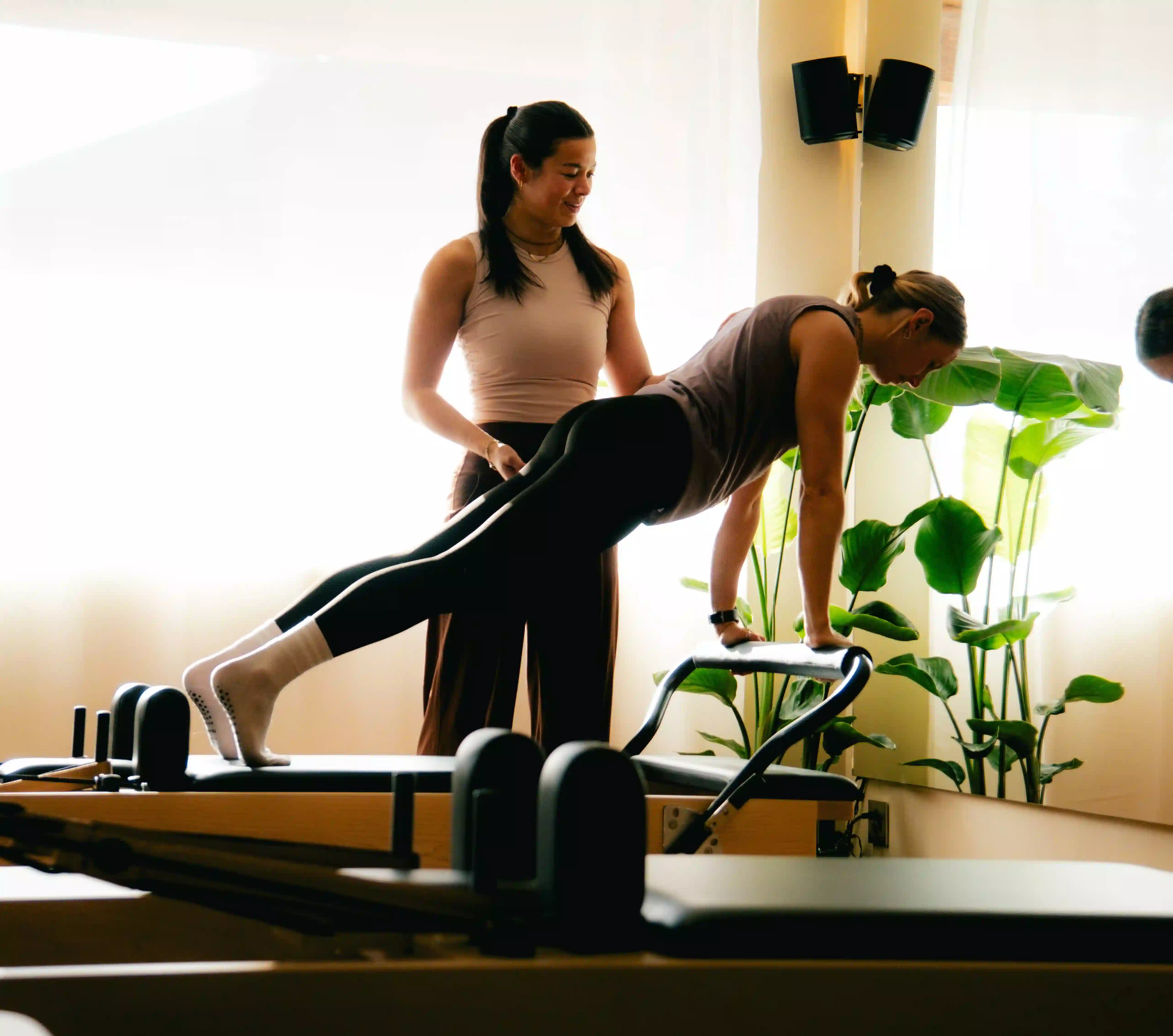 Fitness instructor guides woman wearing black leggings doing plank on Pilates reformer machine.