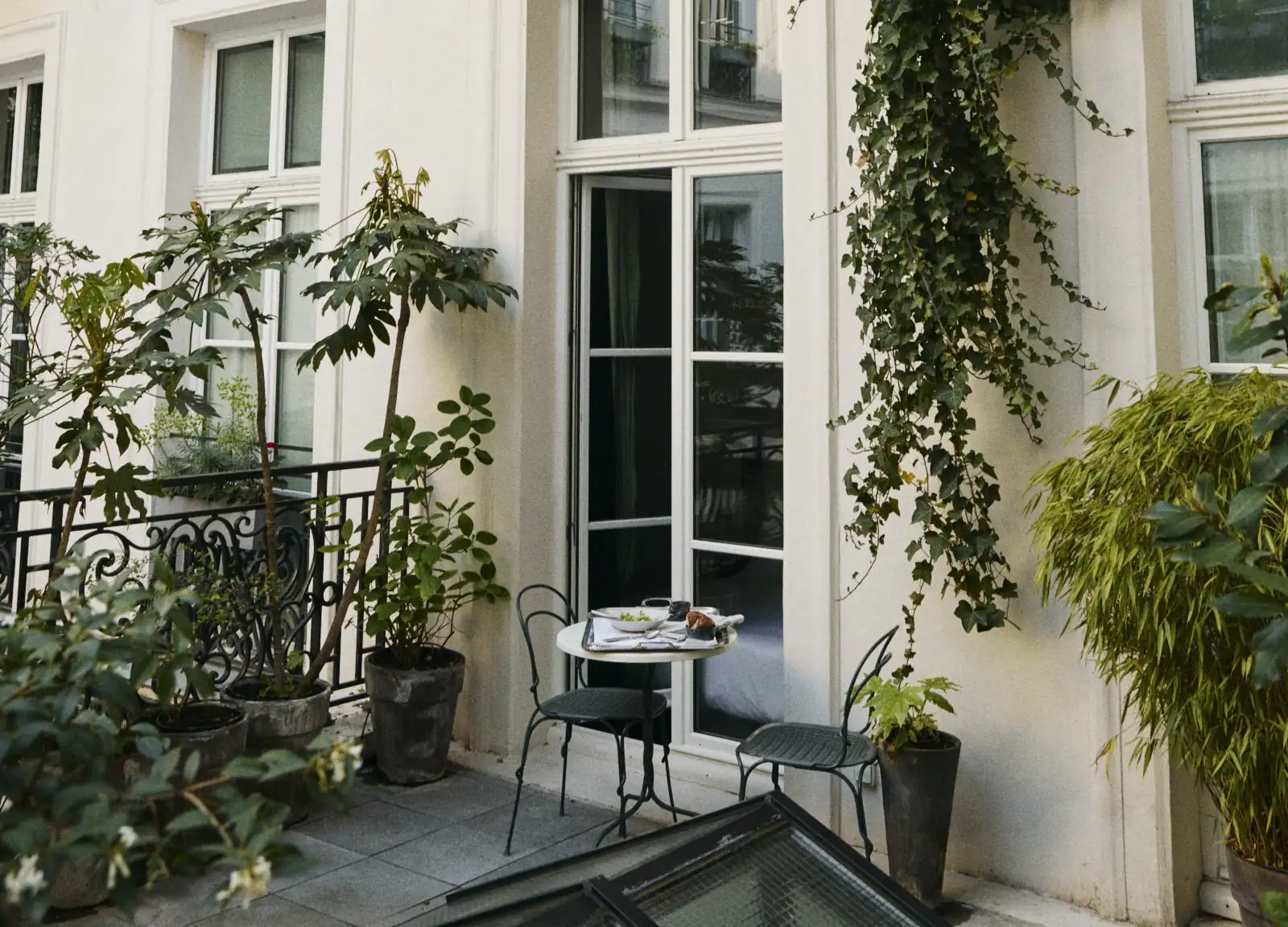 Small balcony with a round table, two chairs, potted plants, and a window leading inside.