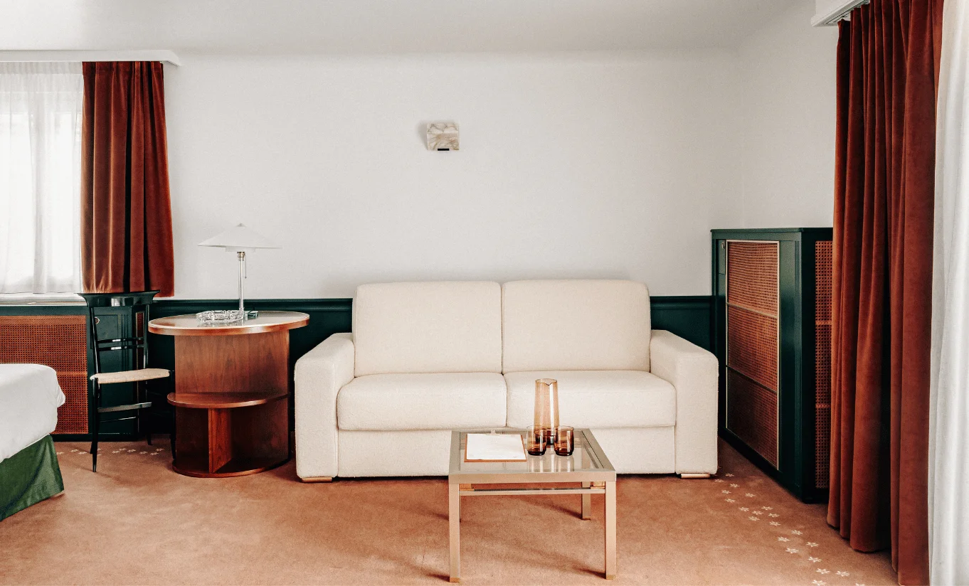 Minimalist hotel room seating area with beige sofa, wooden side table, glass coffee table, and brown curtains.