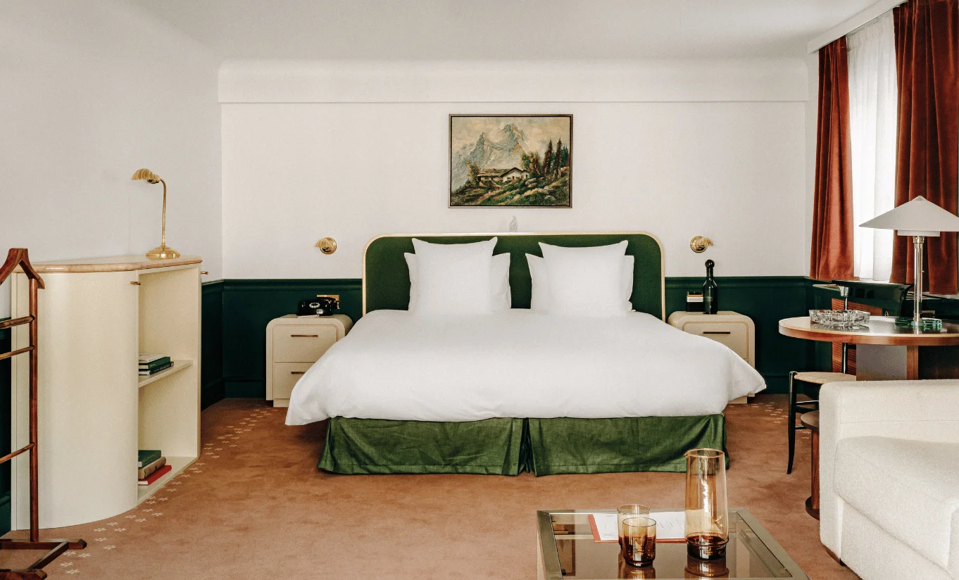 Modern hotel room with green and white king bed, bedside tables, lamp, and a round table near window.