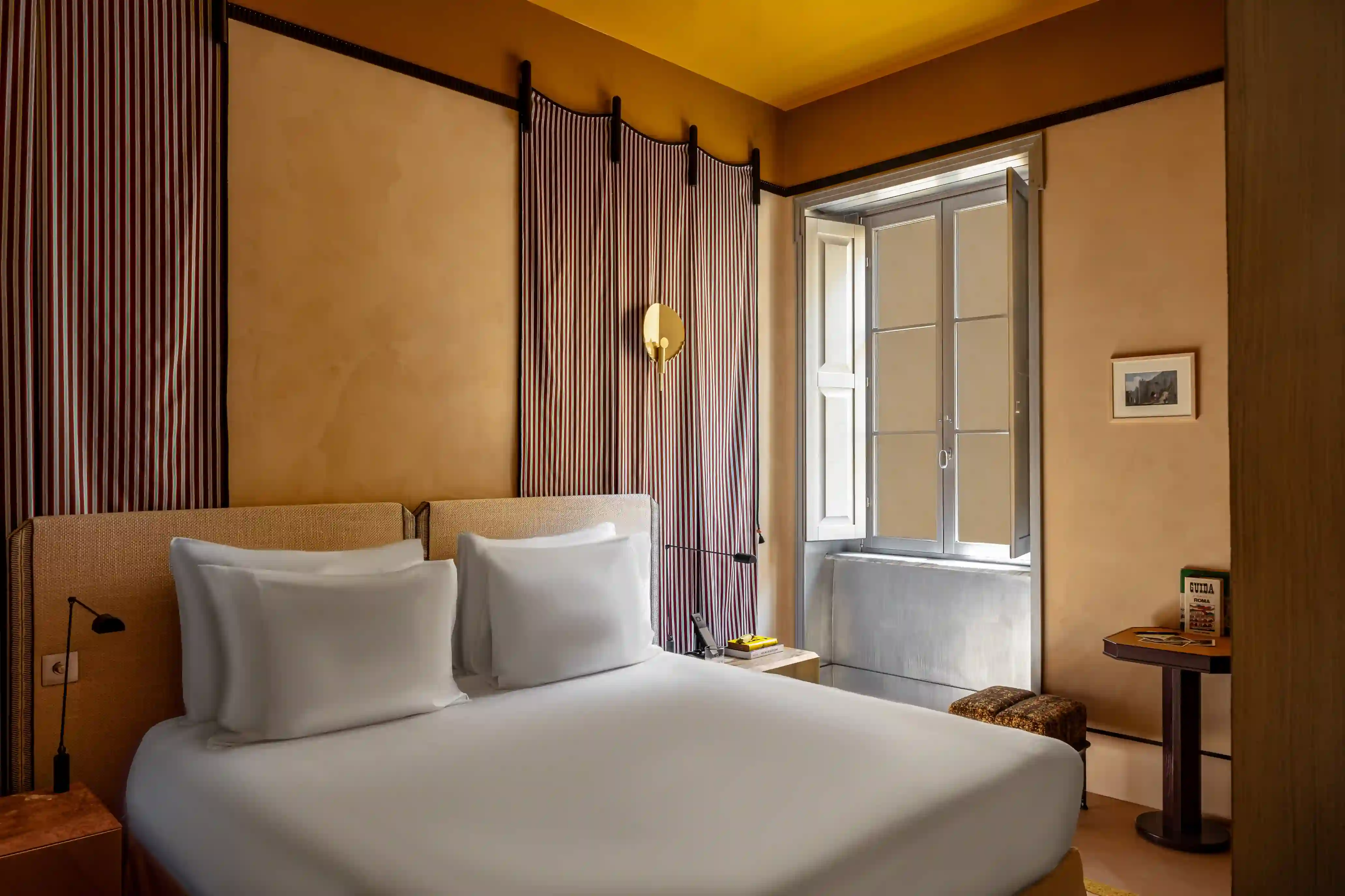 A stylish bedroom interior with white bedding, soft terracotta-colored curtains, and a large window looking out onto an Italian cathedral and square.