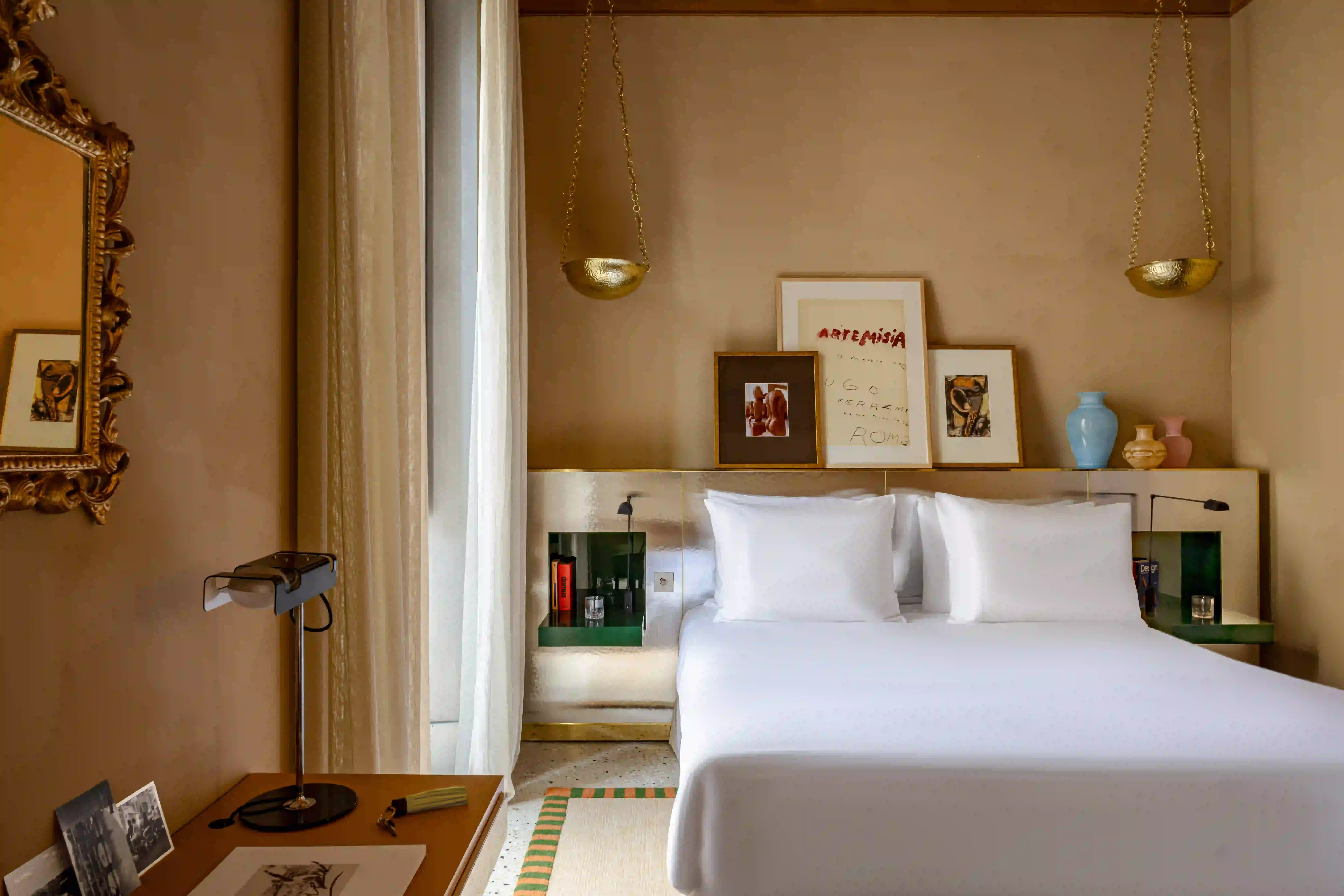 Hotel bedroom with a king bed, framed artwork above the headboard including an Artemisia exhibition poster, hanging brass bowl-shaped sconces, and a vintage mirror by the window.