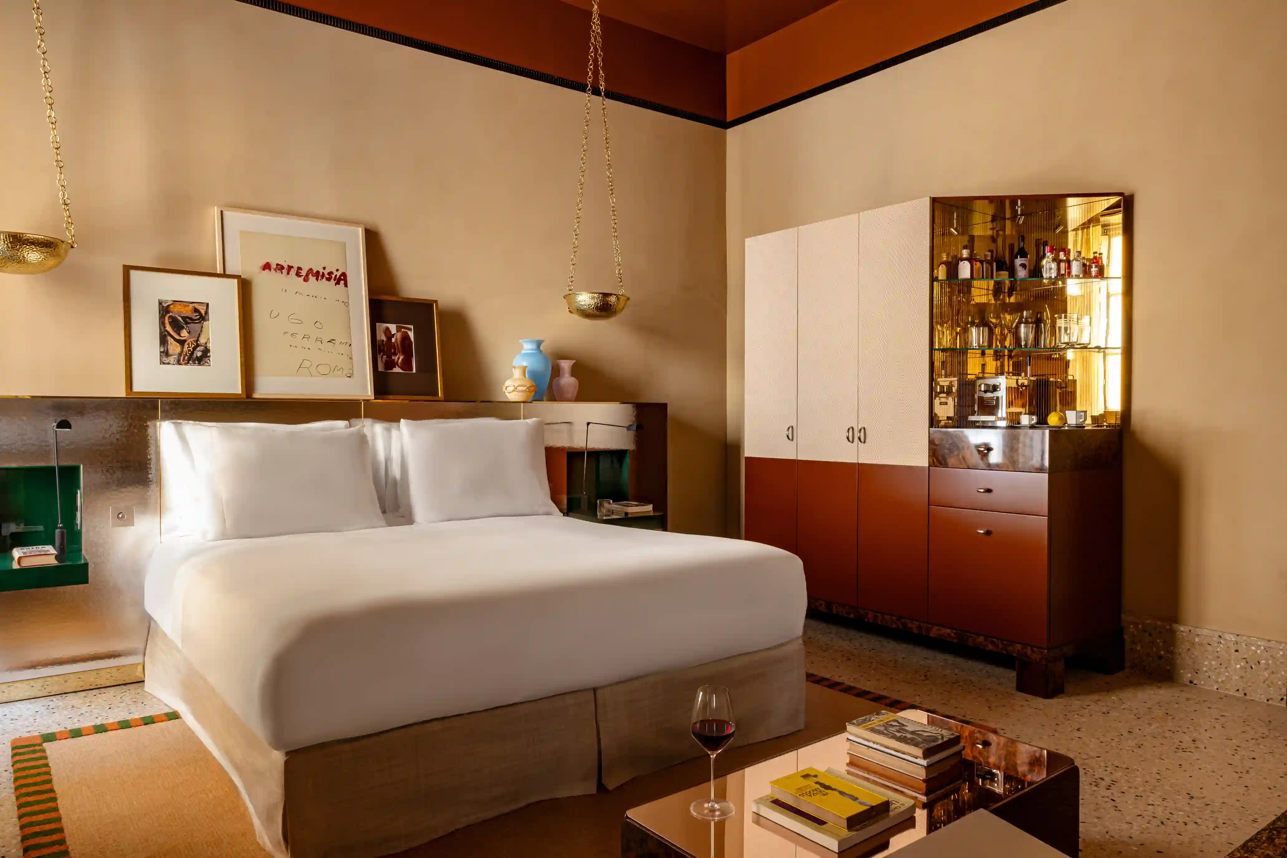 Hotel suite bedroom with a king bed, framed artwork above the headboard, brass hanging sconces, and a two-tone bar cabinet stocked with bottles and glassware.