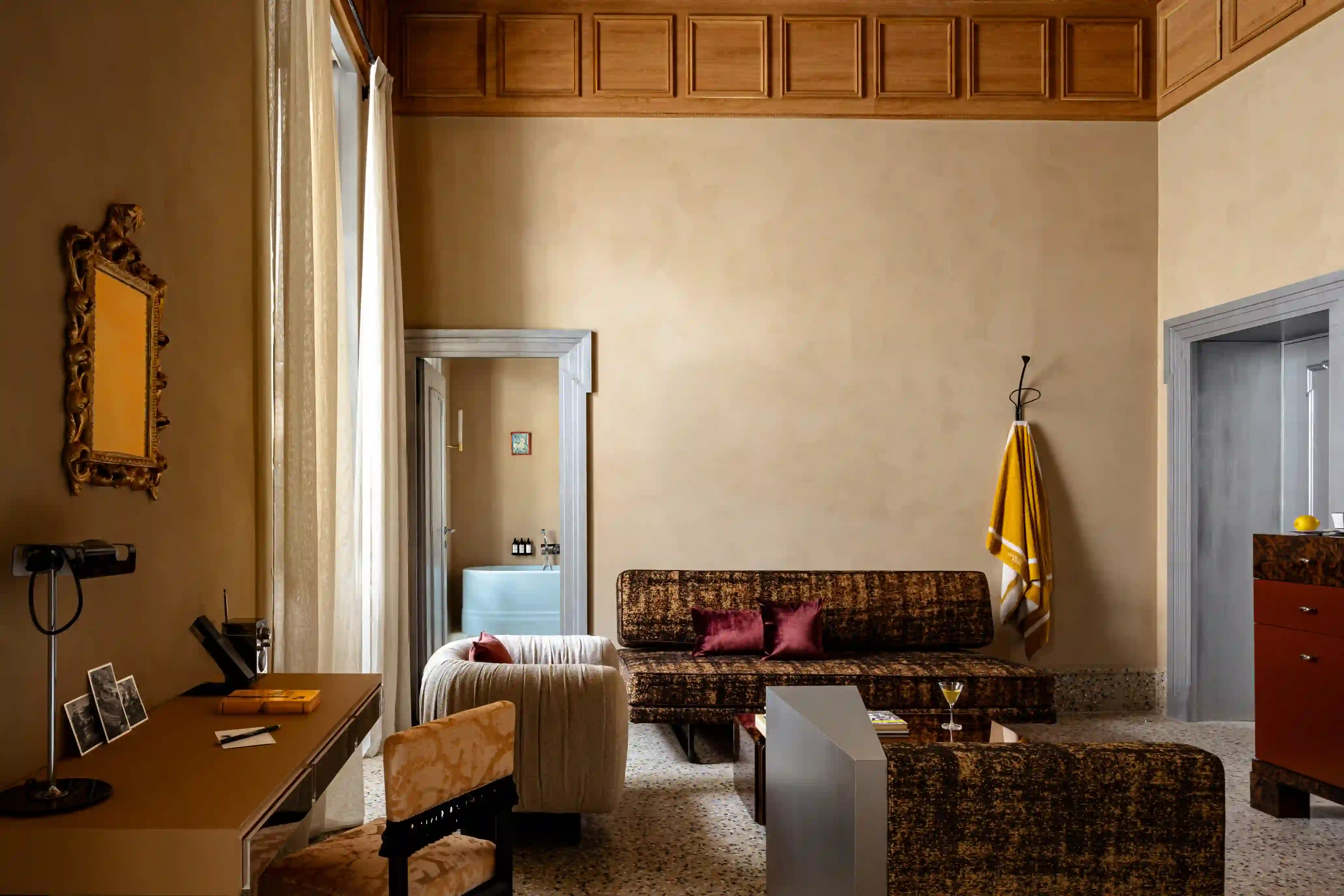 Hotel suite living area with a patterned sofa and burgundy cushions, an ornate gilt mirror, woodpaneled ceiling trim, and a view into an en-suite bathroom with a freestanding tub.
