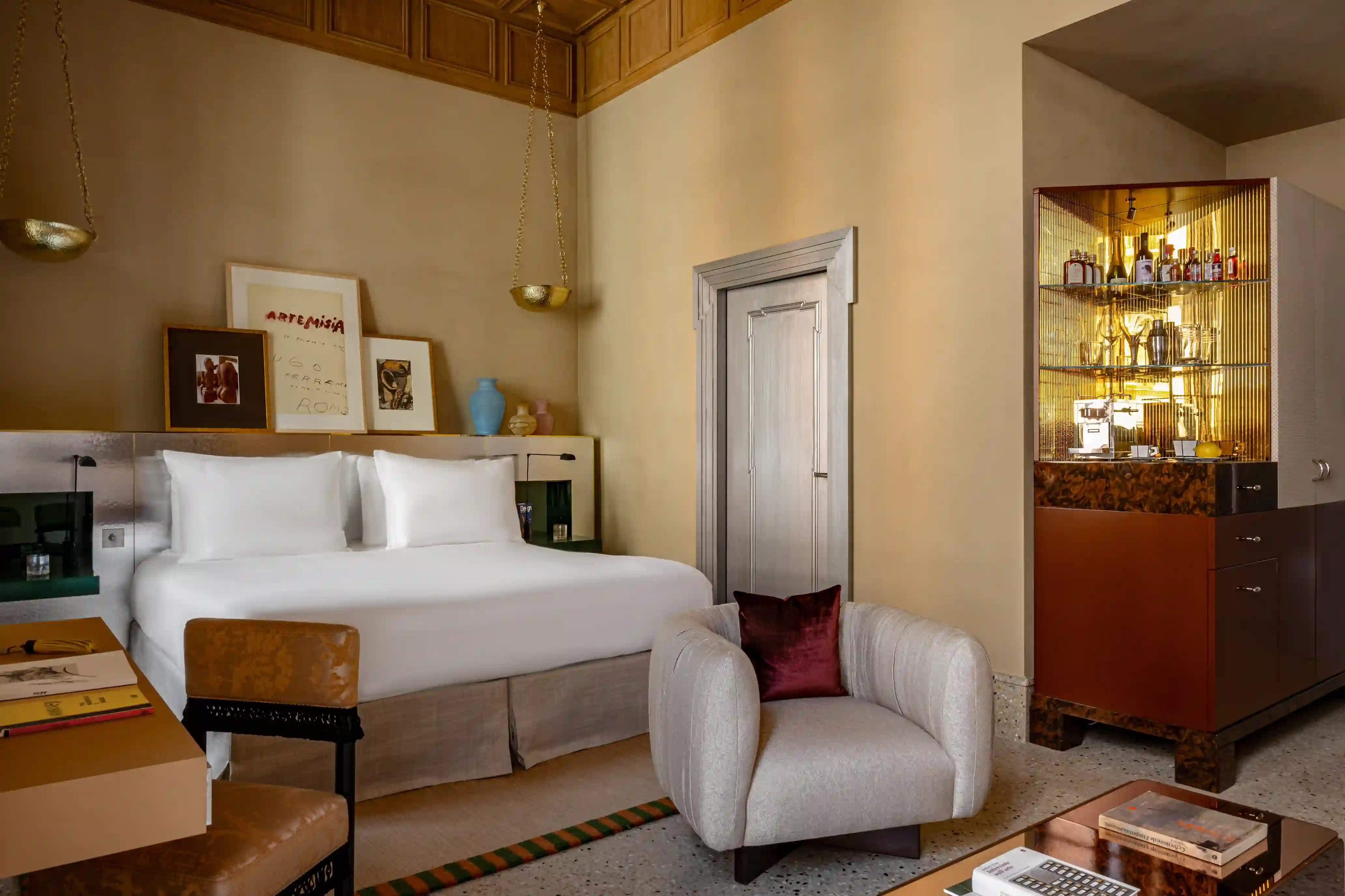Hotel suite bedroom with a king bed, a gallery of framed art above the headboard, a cream armchair with a burgundy cushion, and a glass-fronted bar cabinet.