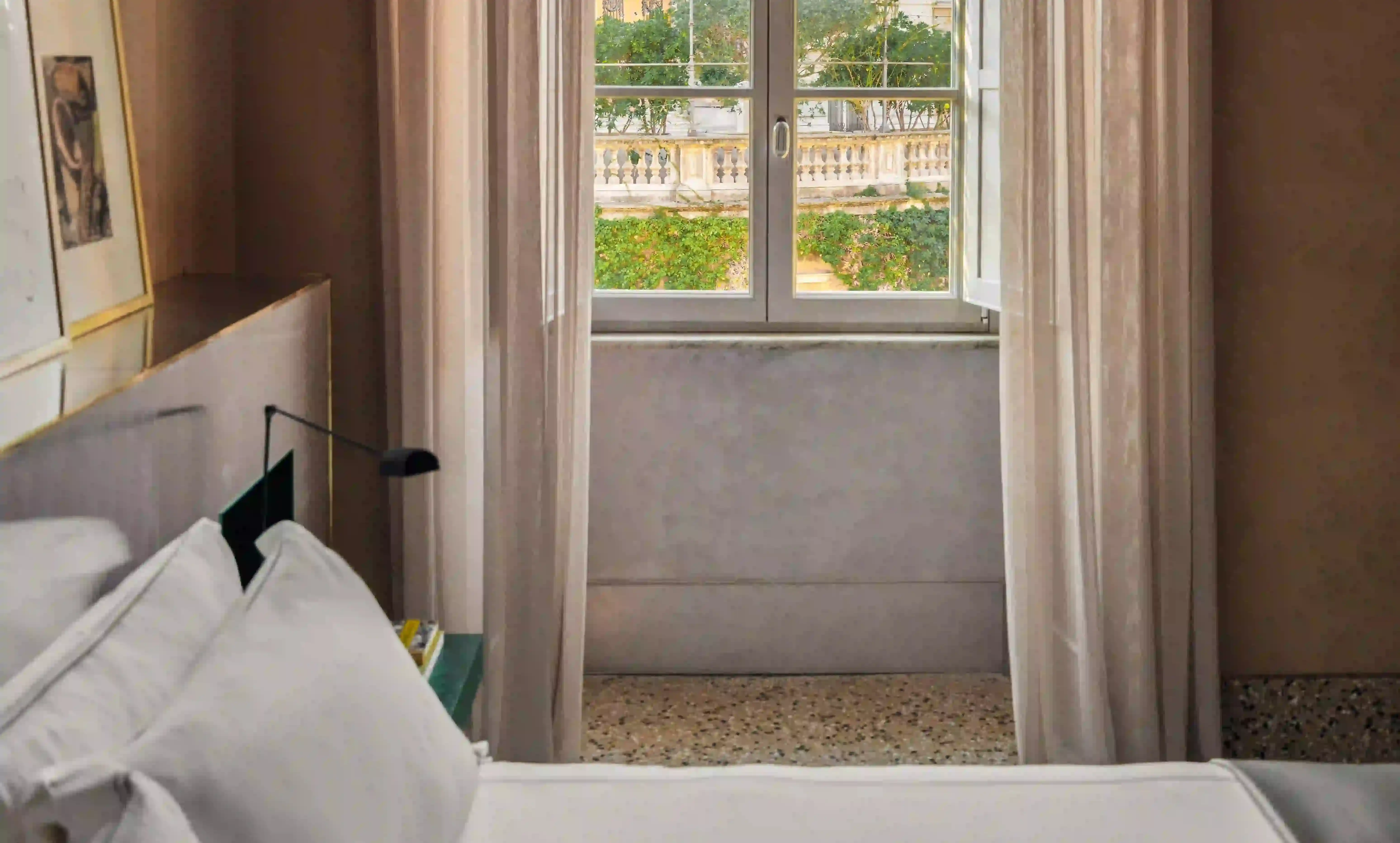 Close view from a hotel bed toward an open window with sheer curtains, overlooking a balustraded terrace and garden greenery