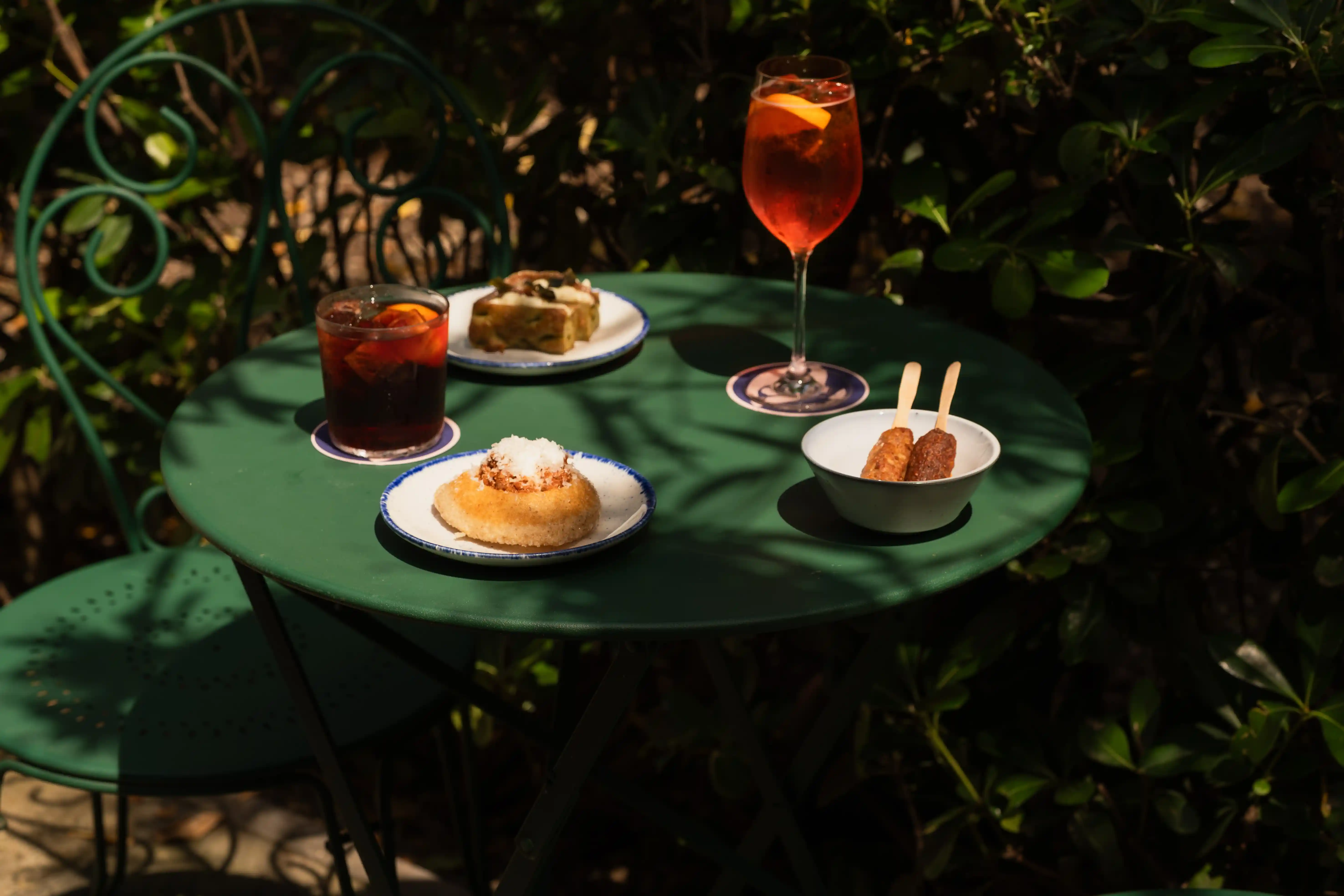A green café table set with an Aperol spritz, a Negroni, and Venetian cicchetti snacks in a garden setting.