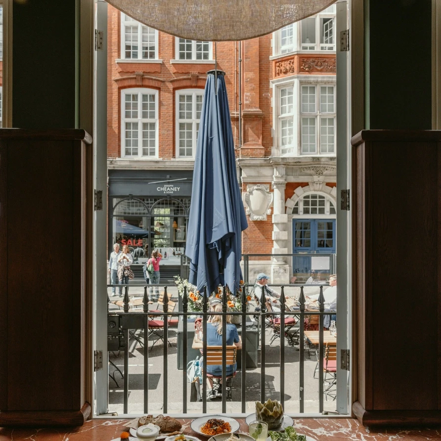 Open window view of sunny terrace with blue parasol and busy Covent Garden street at Henrietta Experimental, London
