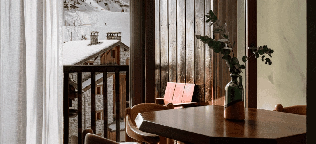 Interno di uno chalet con tavolo e vaso davanti a una finestra con vista su una stazione sciistica innevata