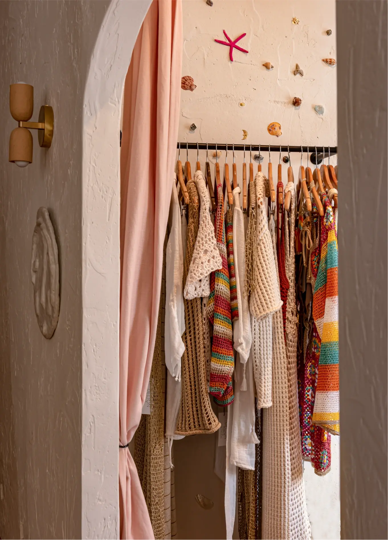 Clothes hang on wooden hangers behind a soft pink curtain in a boutique with seashell wall decor.