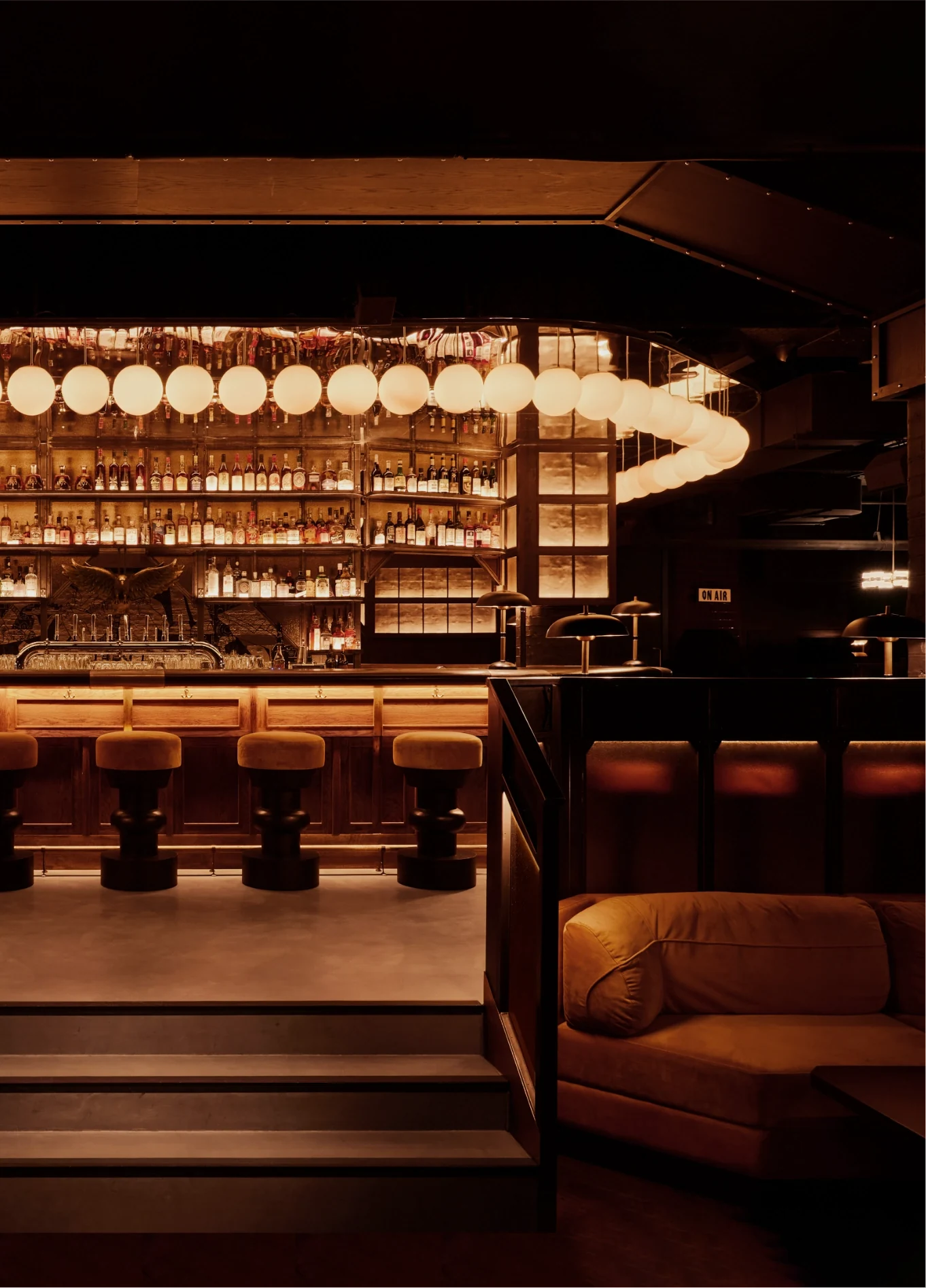 Warmly lit bar interior with stools, shelves of bottles, round hanging lights, and cozy seating area.