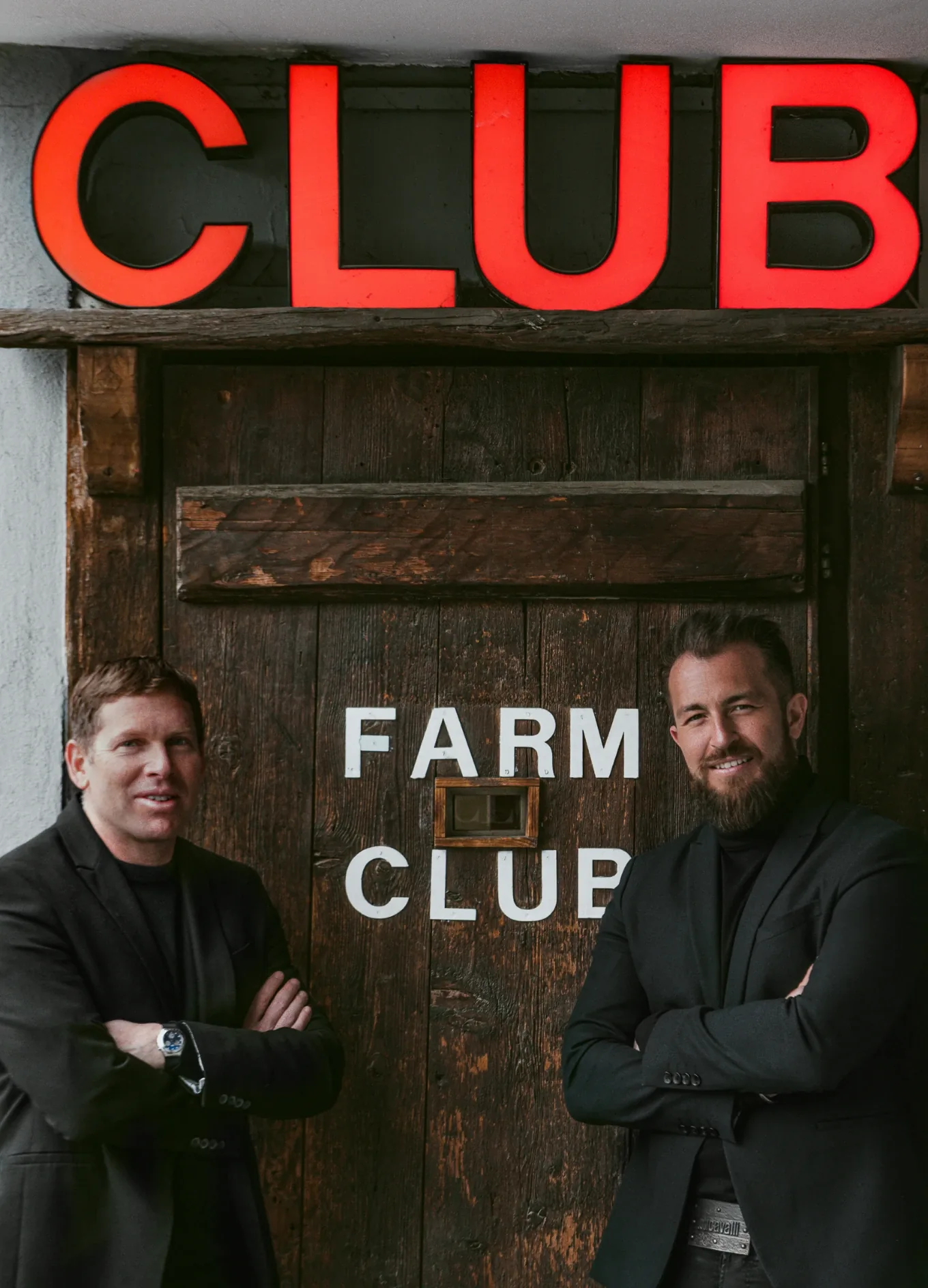 Two men in black jackets stand with arms crossed outside a wooden door reading FARM CLUB.