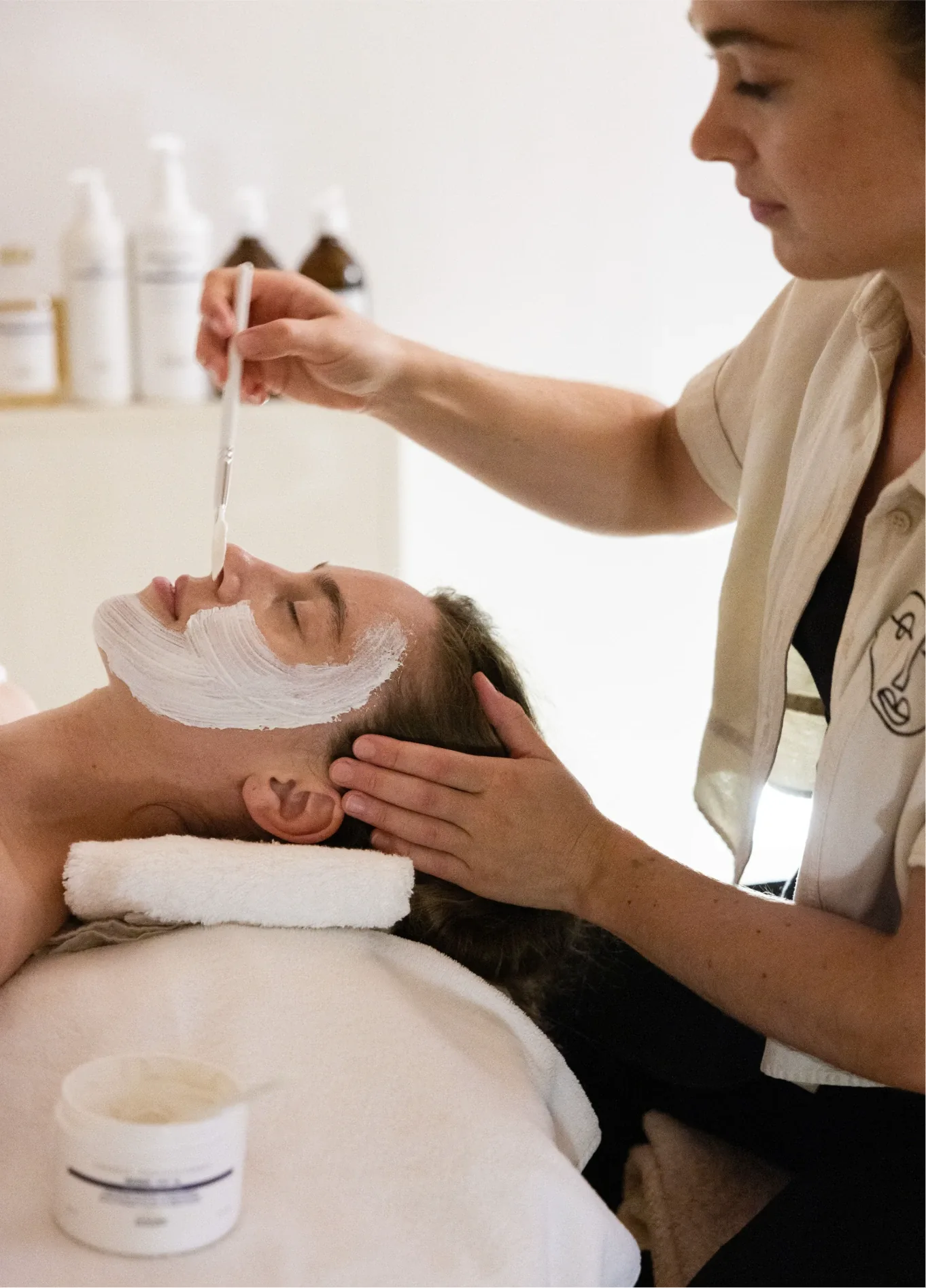 Woman applying a white face mask on a client lying with eyes closed during a spa treatment.