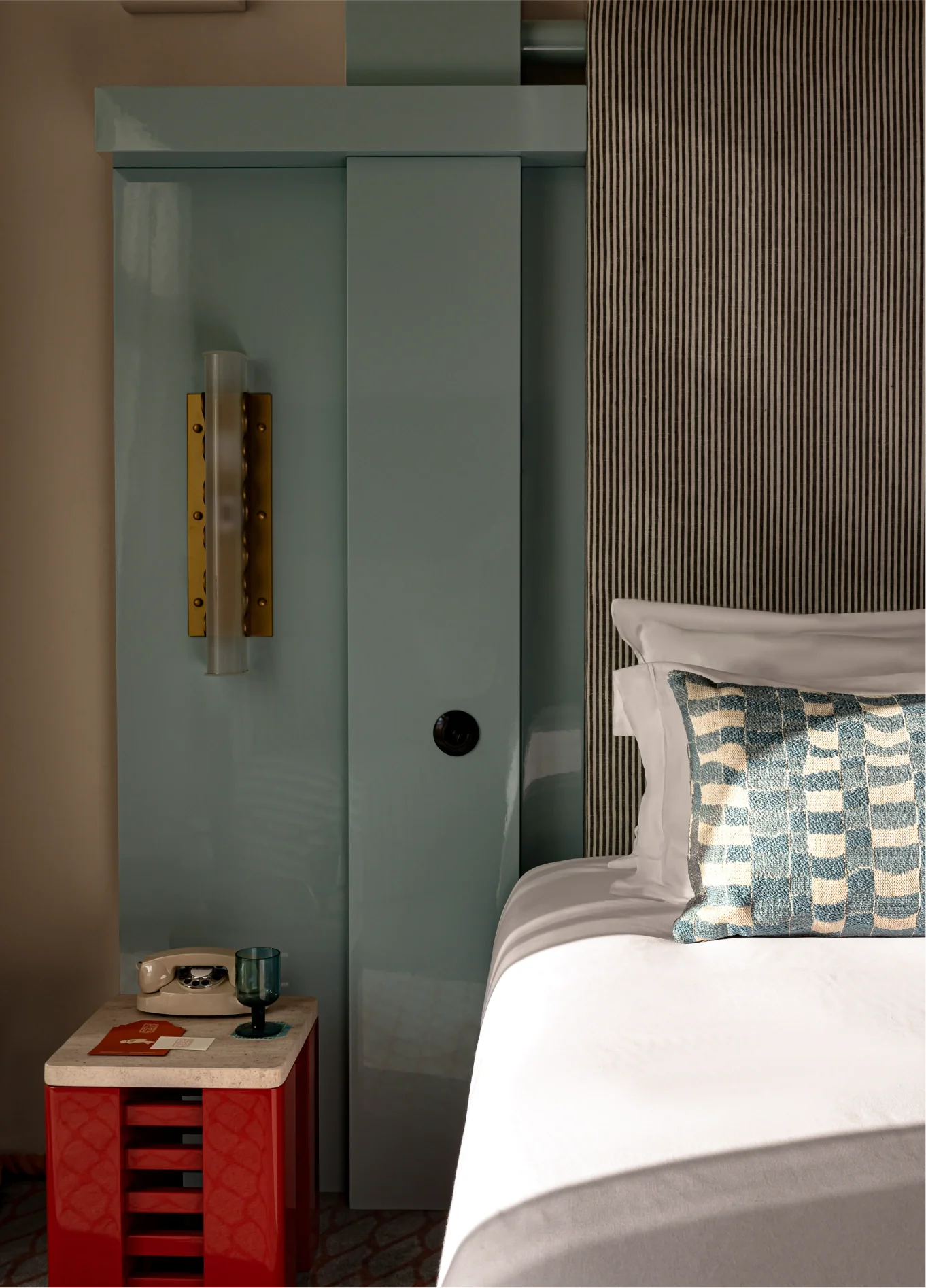 Partial bed with a patterned pillow, a red nightstand holding a vintage phone and glass, beside a glossy blue panel wall.