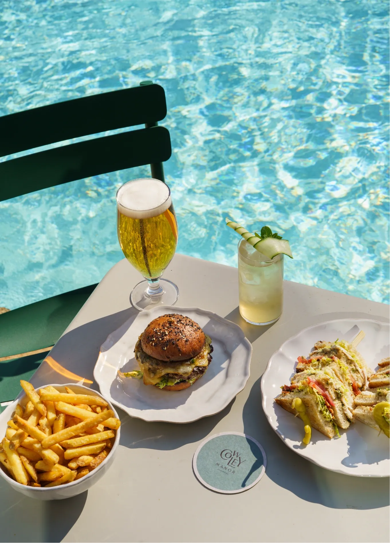 Burger, fries, cocktail, and beer on a table by a clear blue pool with a green chair nearby.