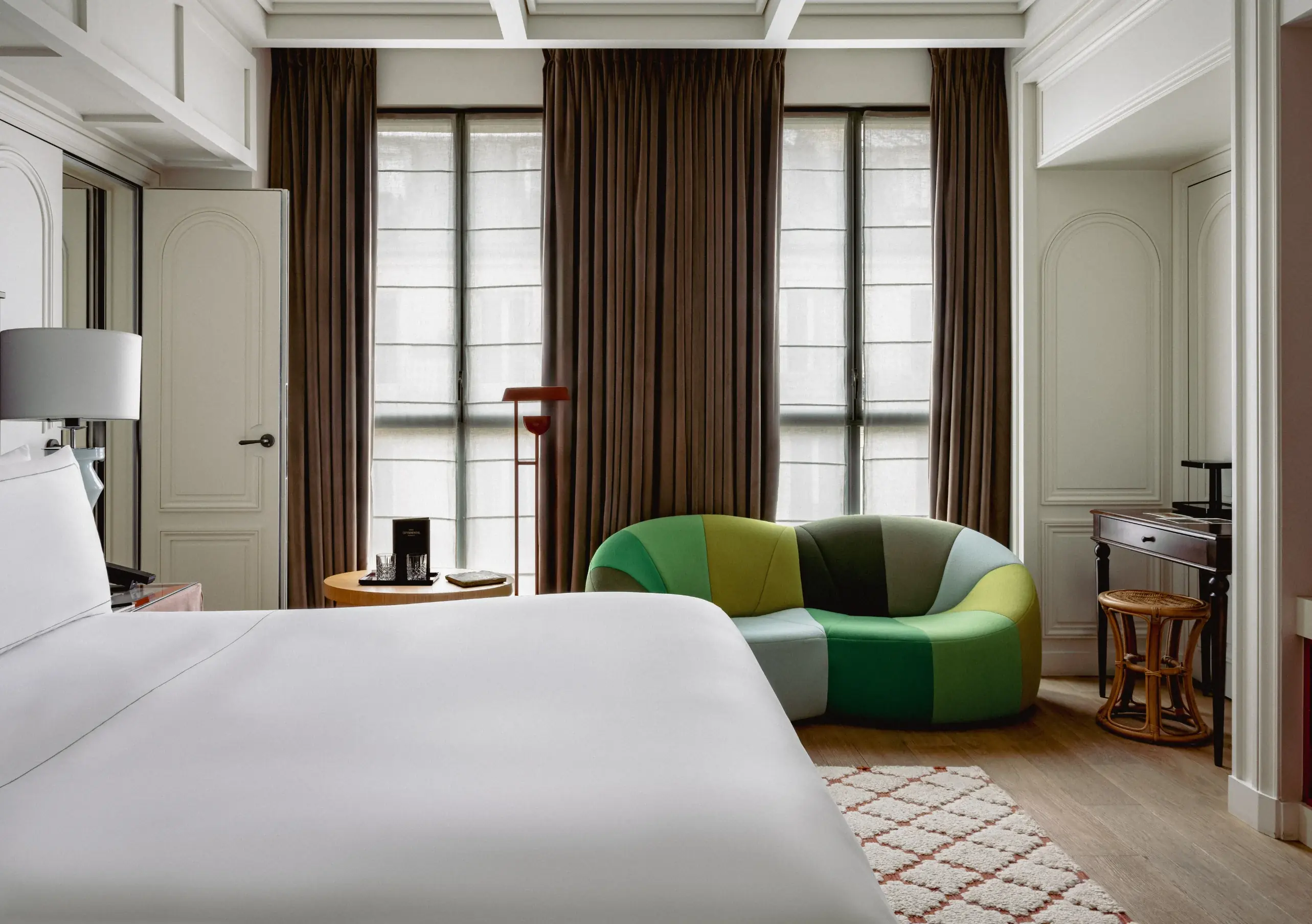 Hotel room with a large white bed, green patchwork sofa, round table, and floor-to-ceiling windows.