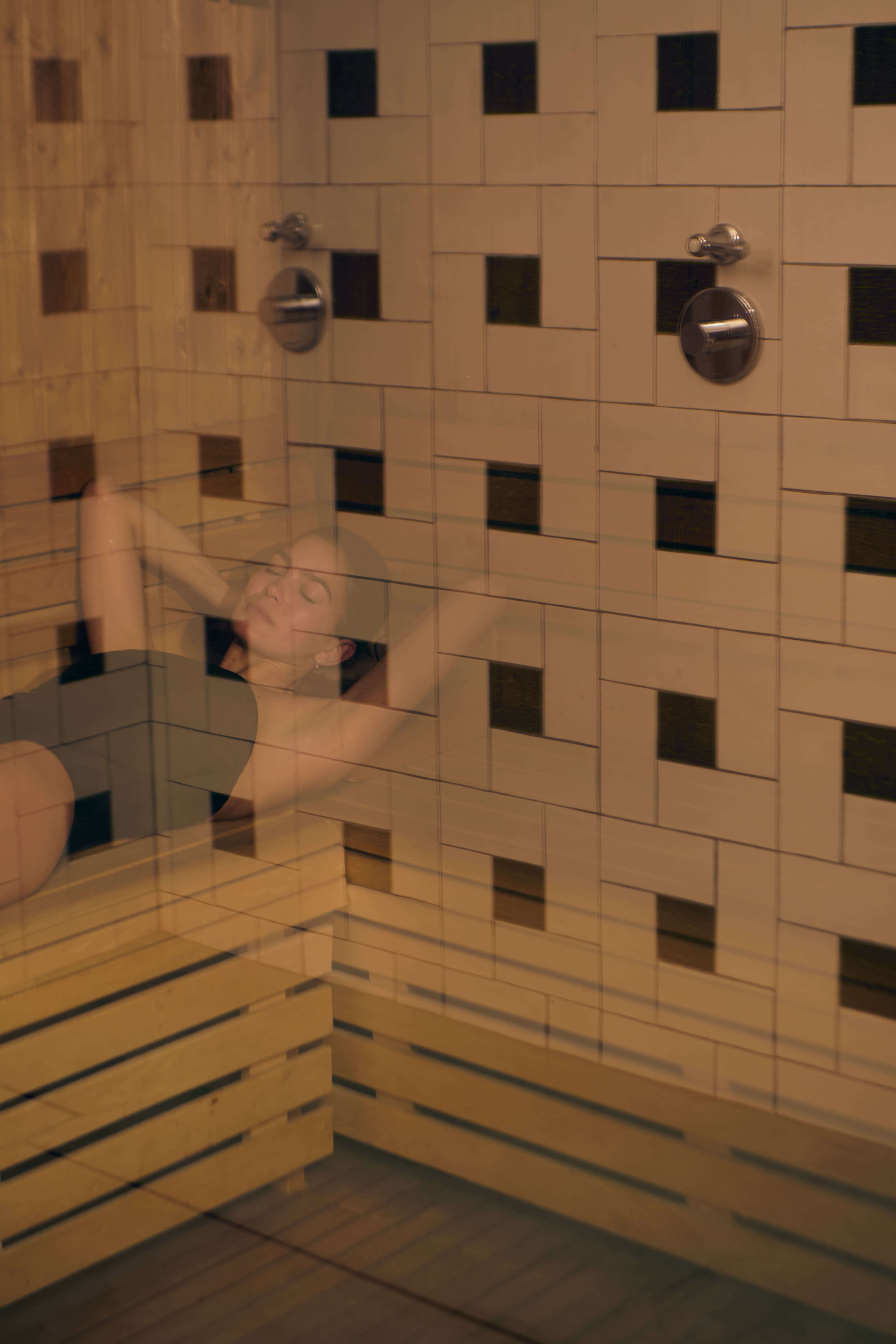 Woman in black swimsuit relaxing on a wooden bench in a sauna with tiled walls.