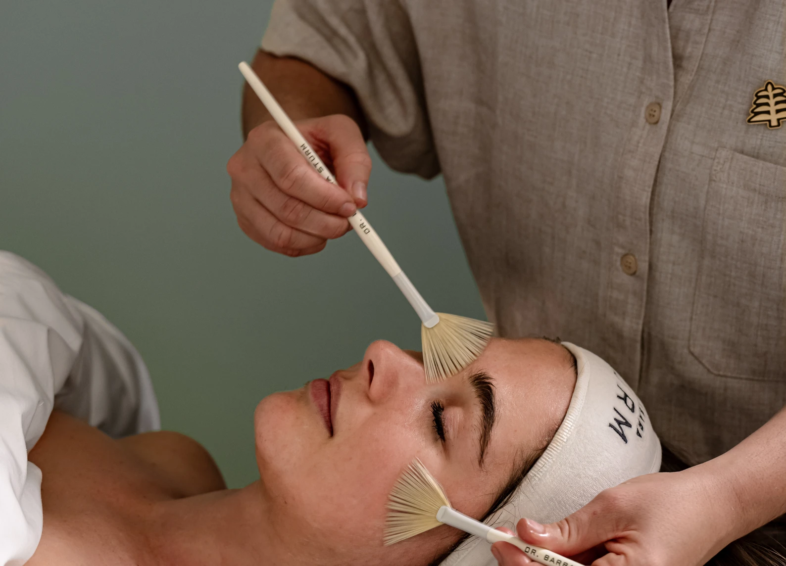 Person receiving facial treatment with two fan brushes on closed eyes and cheek in a spa setting.