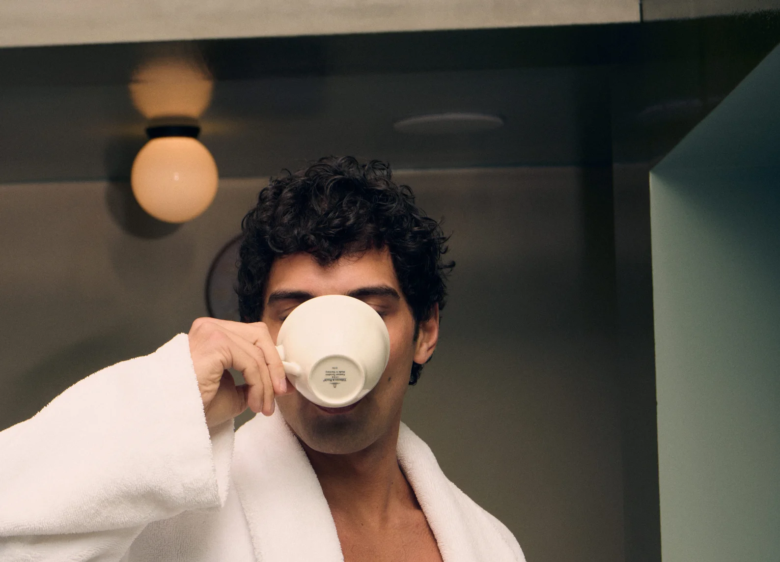 Man with curly hair in a white robe drinking from a white ceramic cup indoors.