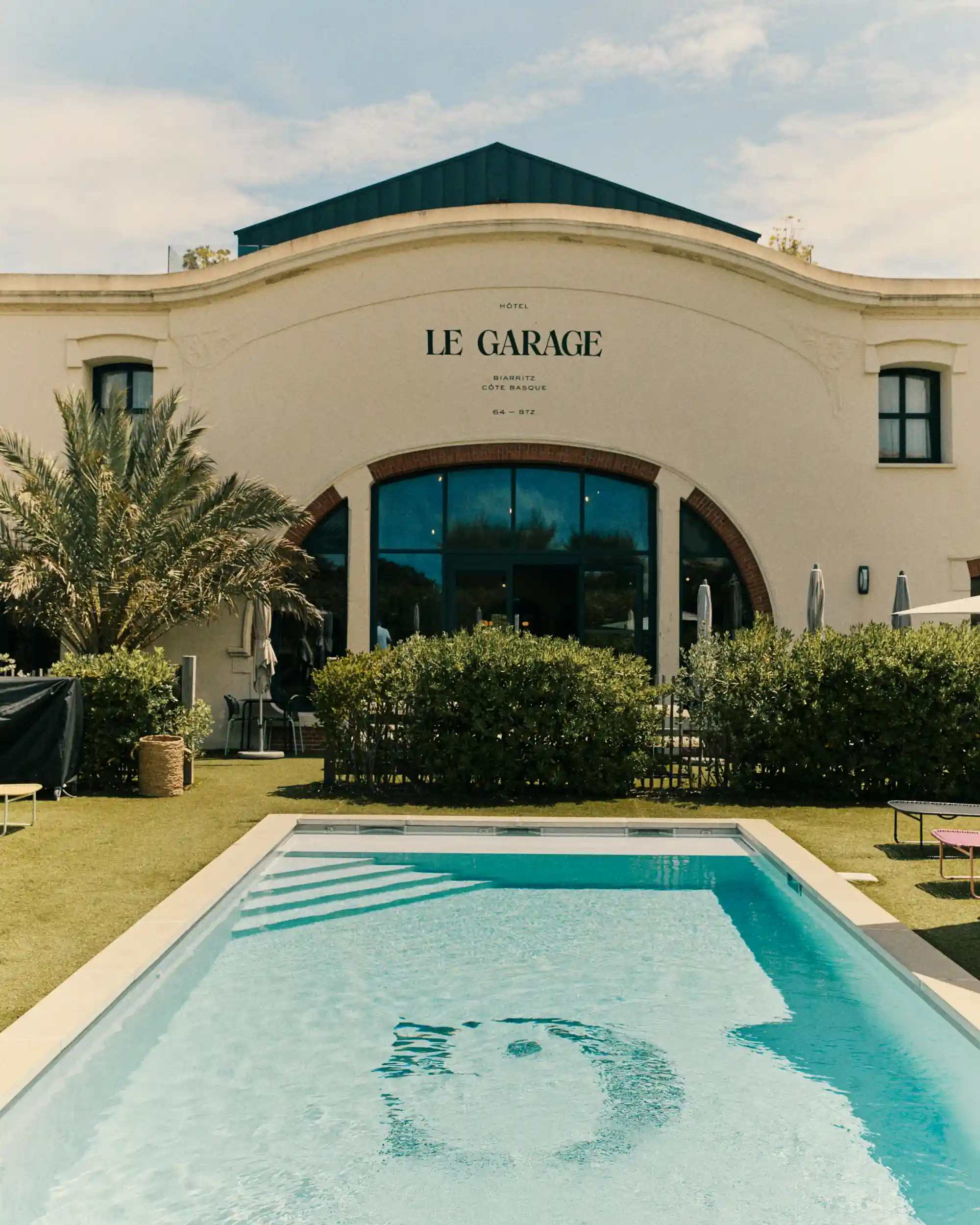 Outdoor pool in front of the white arched facade of Hotel Le Garage in Biarritz under clear sky.
