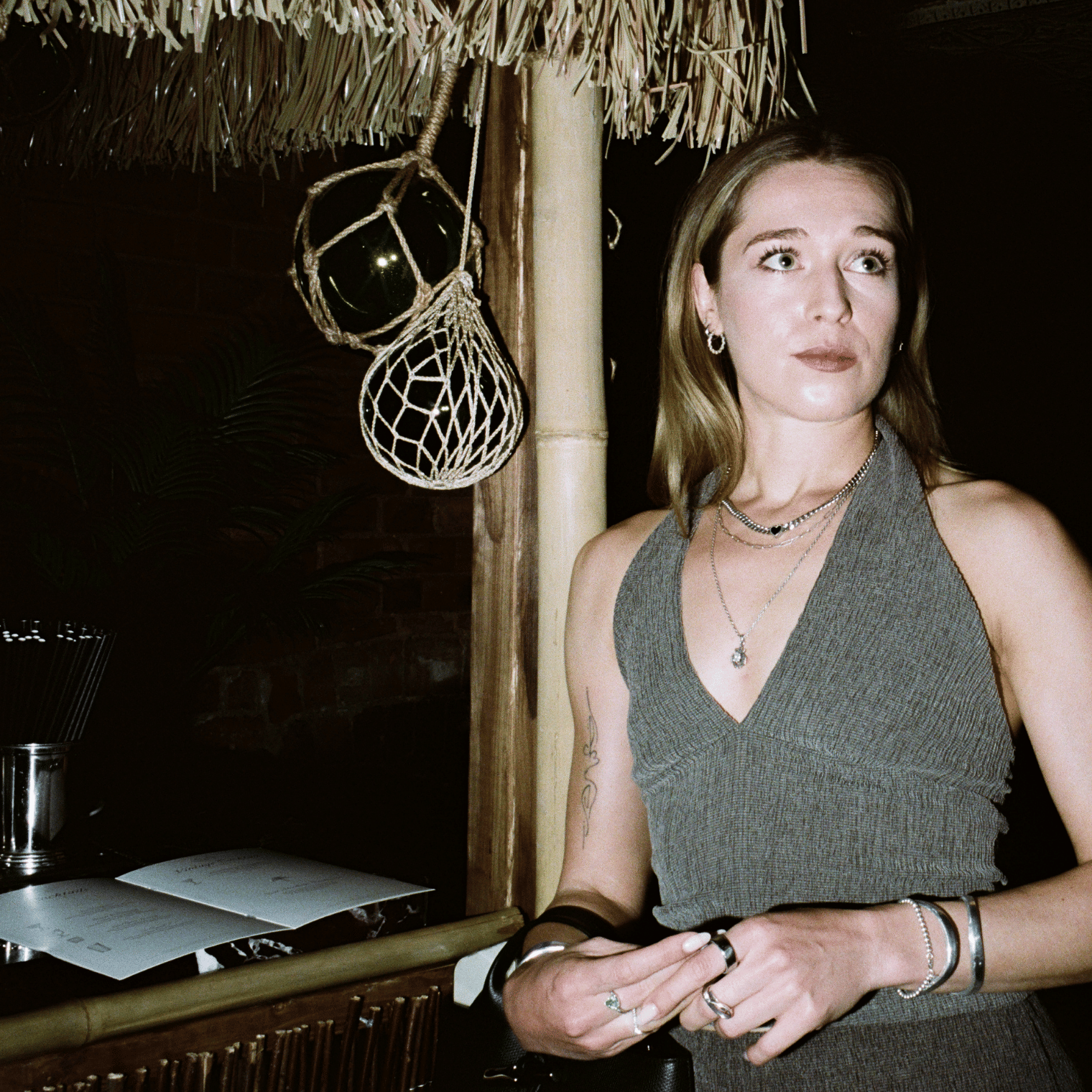 Woman with layered necklaces and earrings stands near bamboo post under a thatched roof at night.