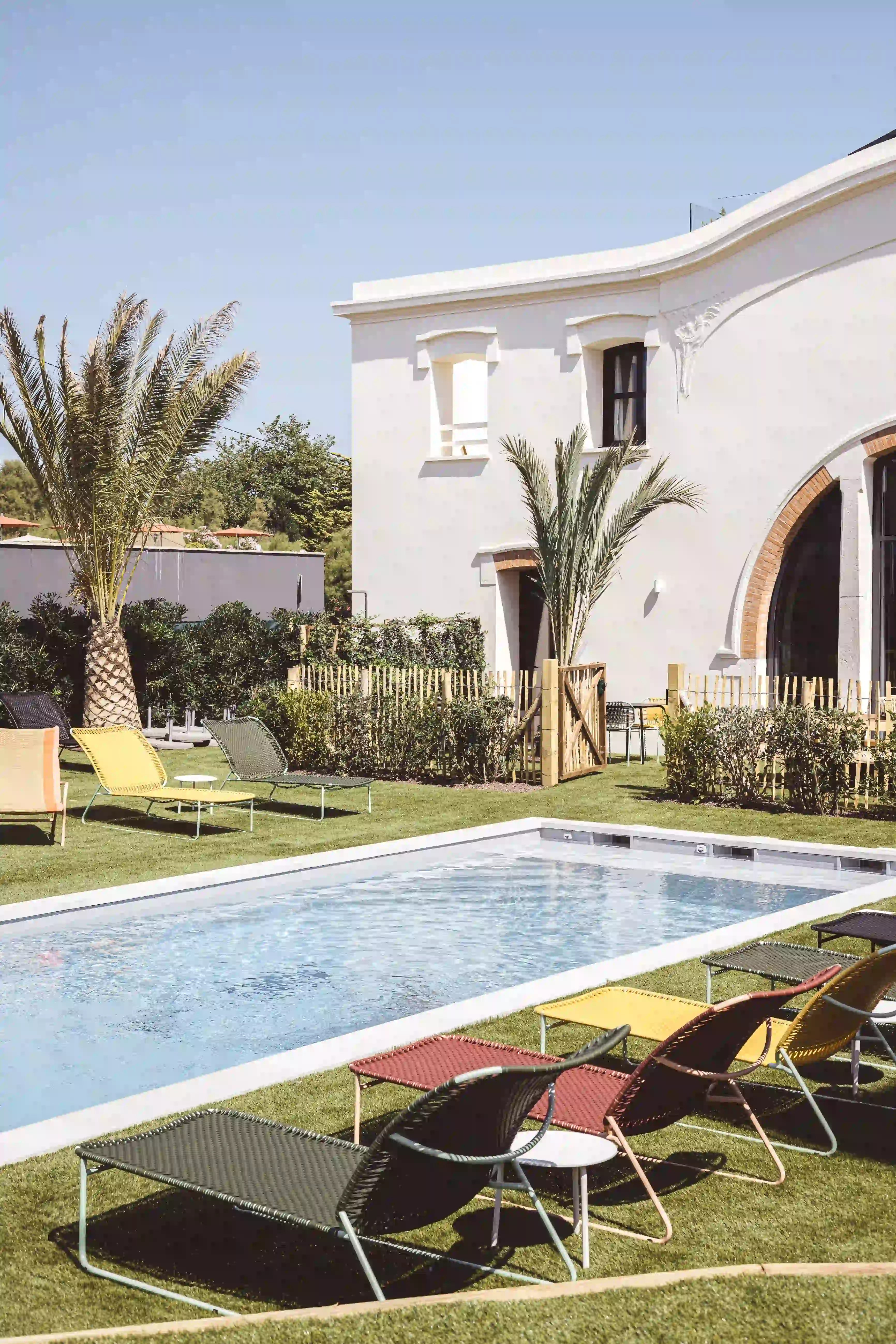 Outdoor pool surrounded by colorful lounge chairs and palm trees beside a white building with arches.