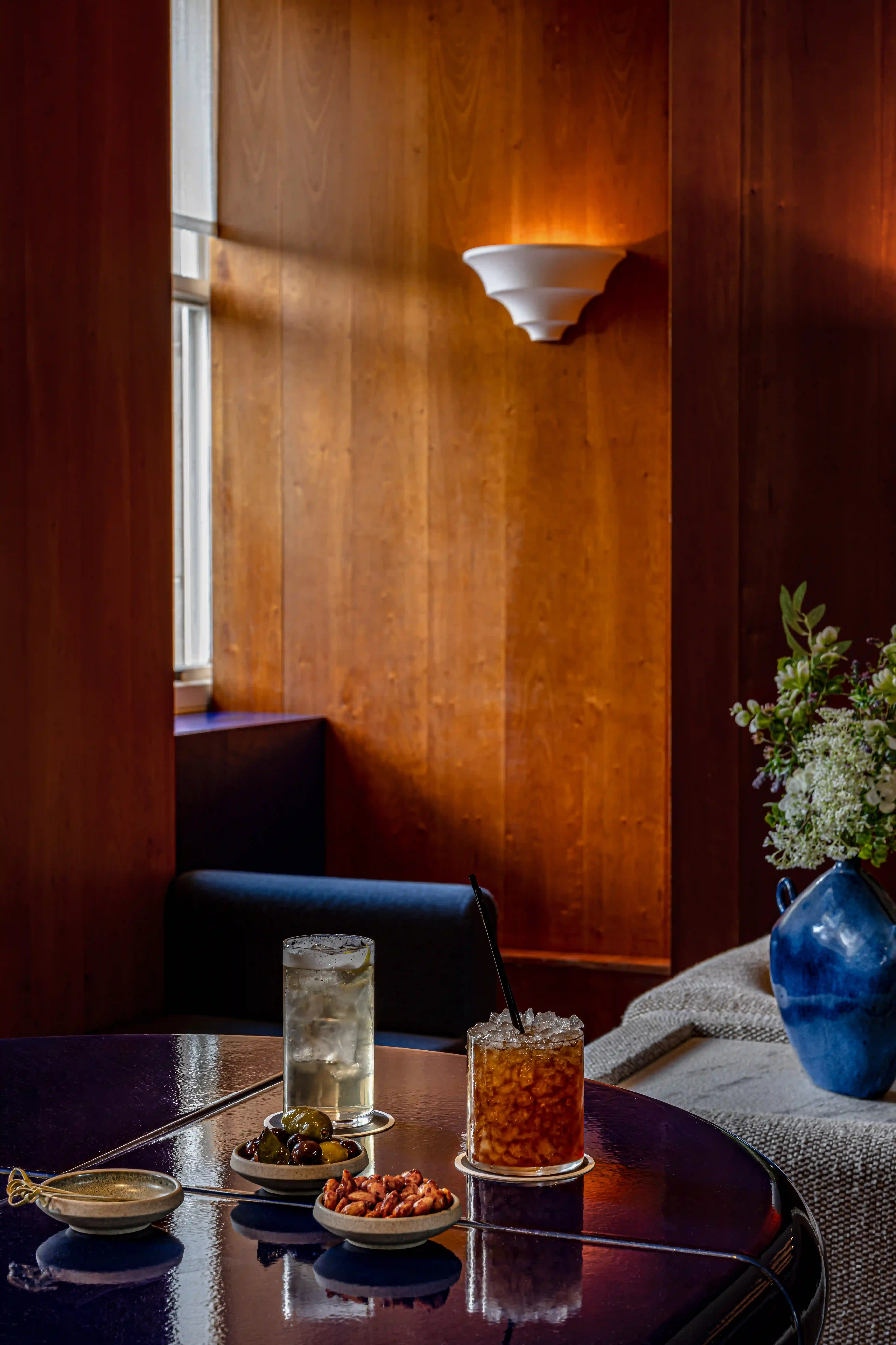 Modern lounge with textured armchairs, a dark blue vase of white flowers, wood paneled walls, and soft lighting.