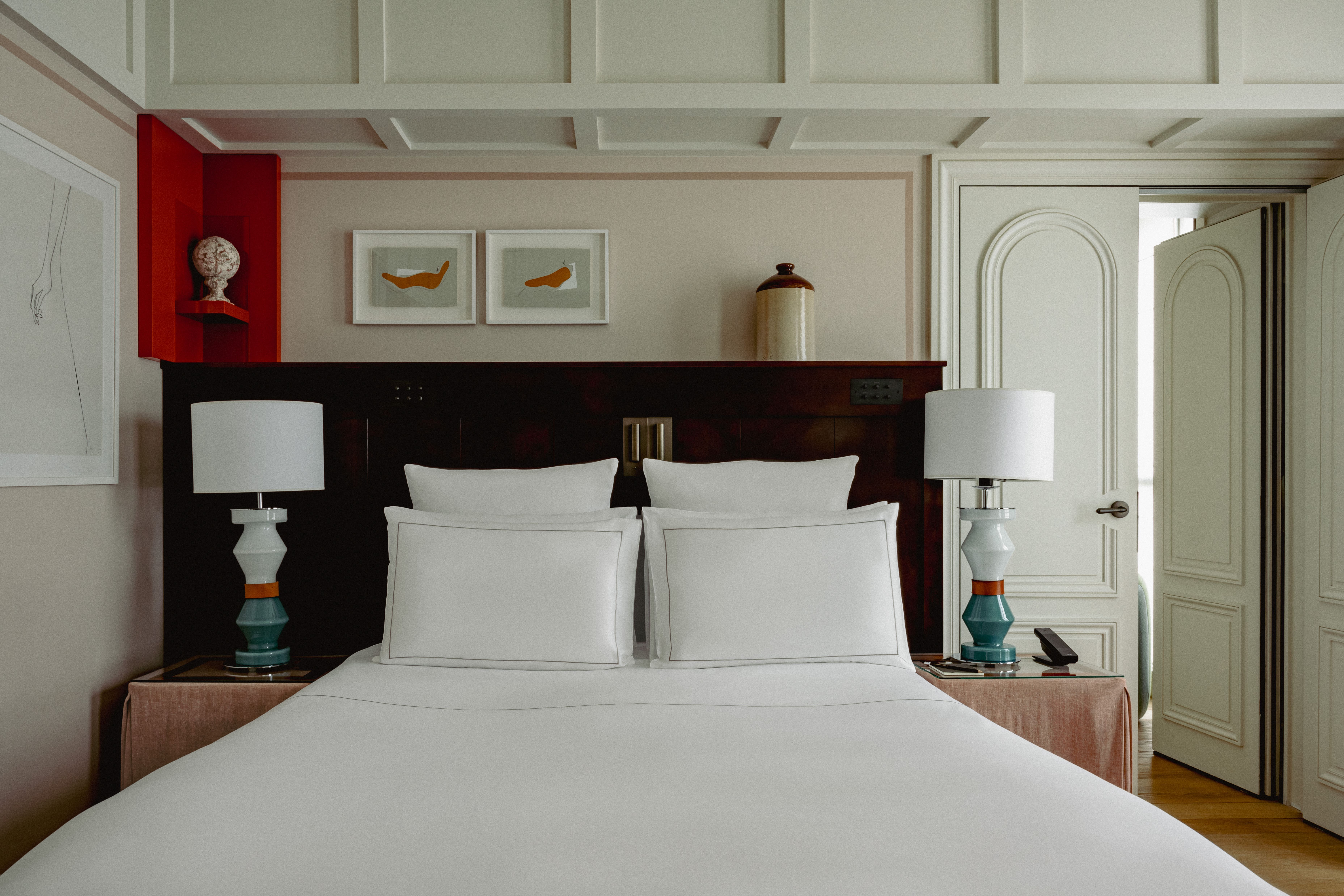 Hotel room interior showing a neatly made bed with white linens and a wooden headboard with a shelf.