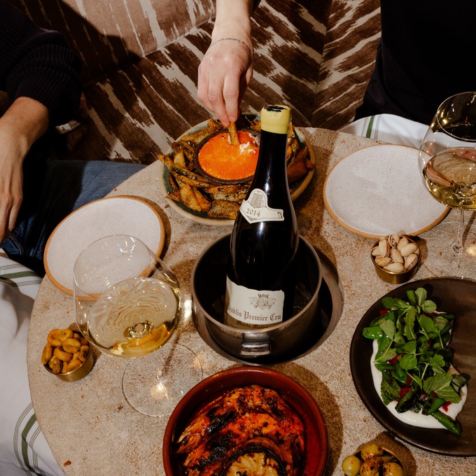 A hand reaching for a potato wedge dipped in orange roe, on a shared table set with wine, grilled seafood, and small plates at a restaurant.