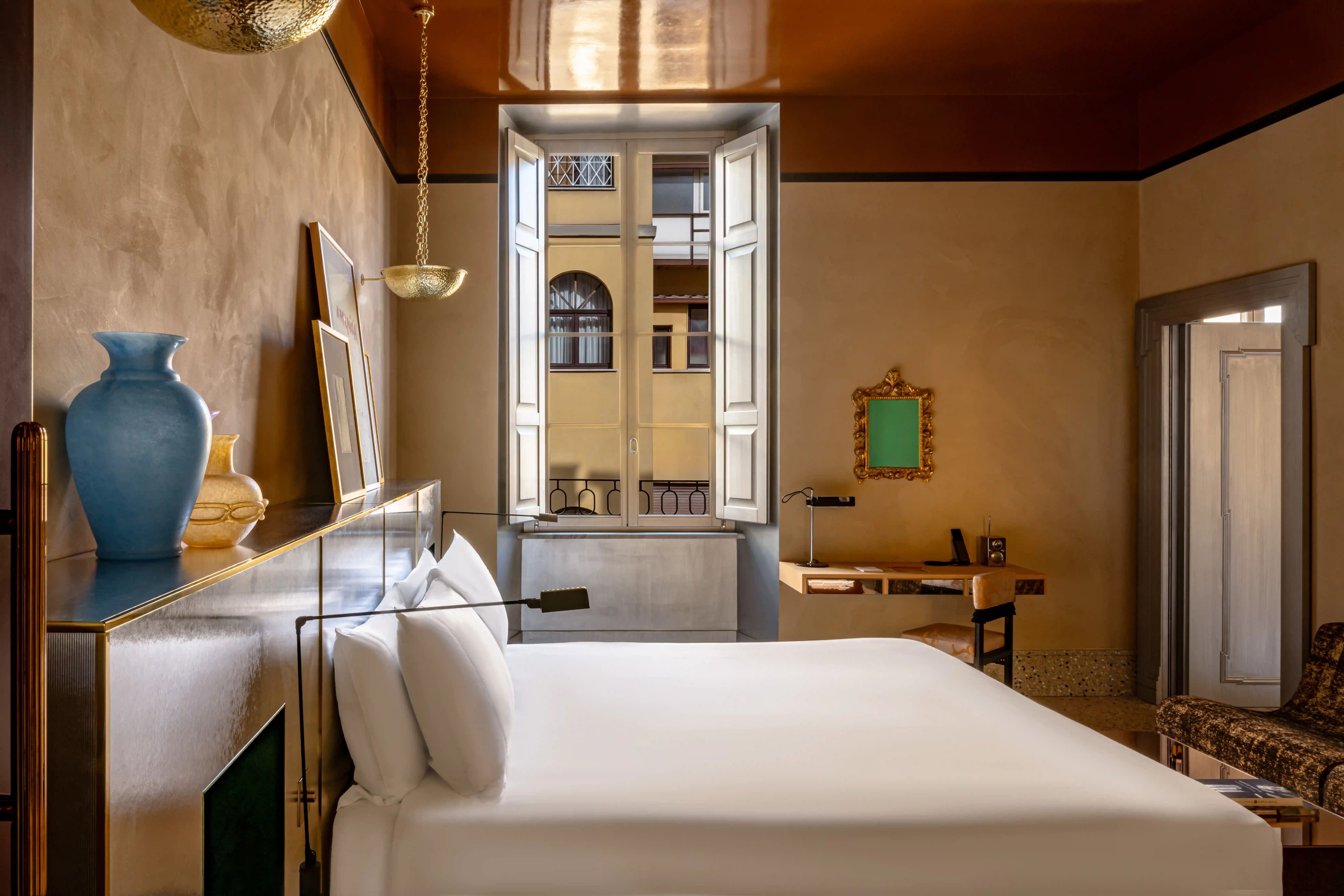 Hotel bedroom with a king bed, framed artwork above the headboard including an Artemisia exhibition poster, hanging brass bowl-shaped sconces, and a vintage mirror by the window.