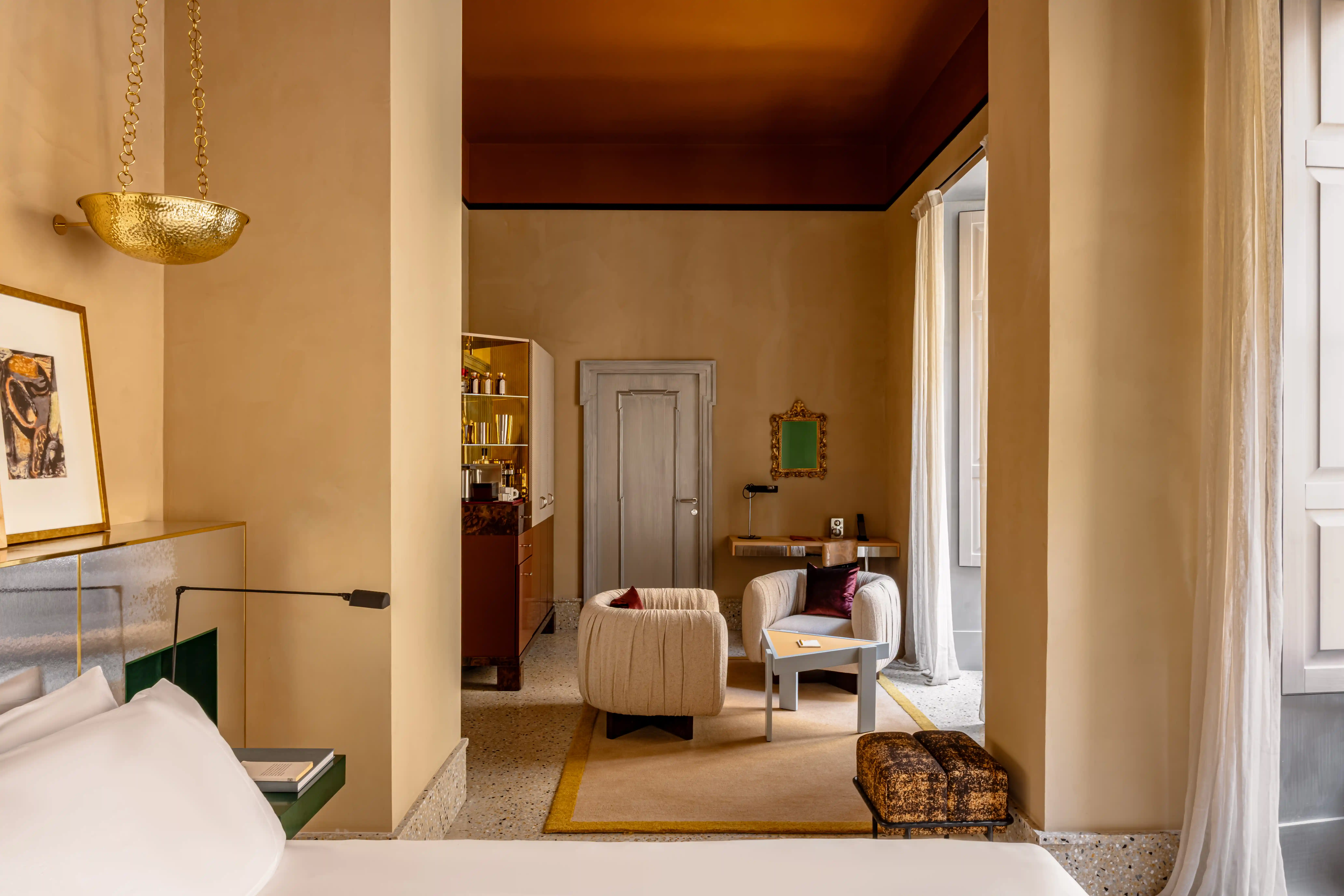 Hotel suite bedroom with a king bed, a gallery of framed art above the headboard, a cream armchair with a burgundy cushion, and a glass-fronted bar cabinet.