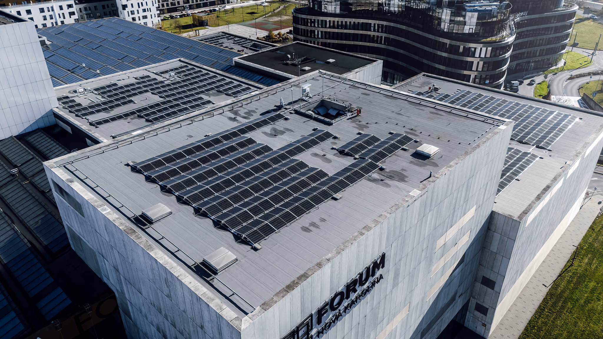 Aerial view of building rooftops covered with solar panels on a sunny day in an urban area with modern buildings and green spaces around.