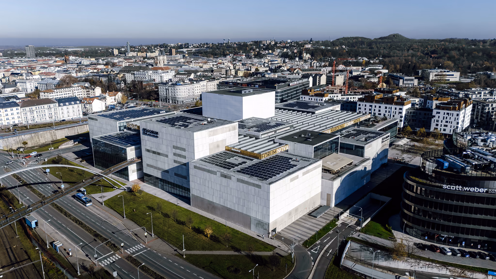 Aerial view of a large modern building with solar panels on the roof, labeled Forum Nova, surrounded by roads and a cityscape with residential and commercial buildings, and greenery in the background under a clear sky.