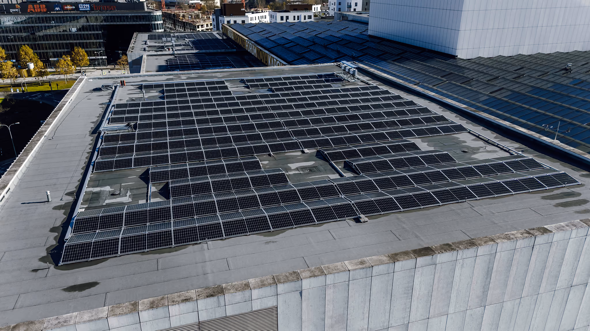 Aerial view of a large flat rooftop covered with numerous solar panels on a white building, with other buildings and trees visible in the background.